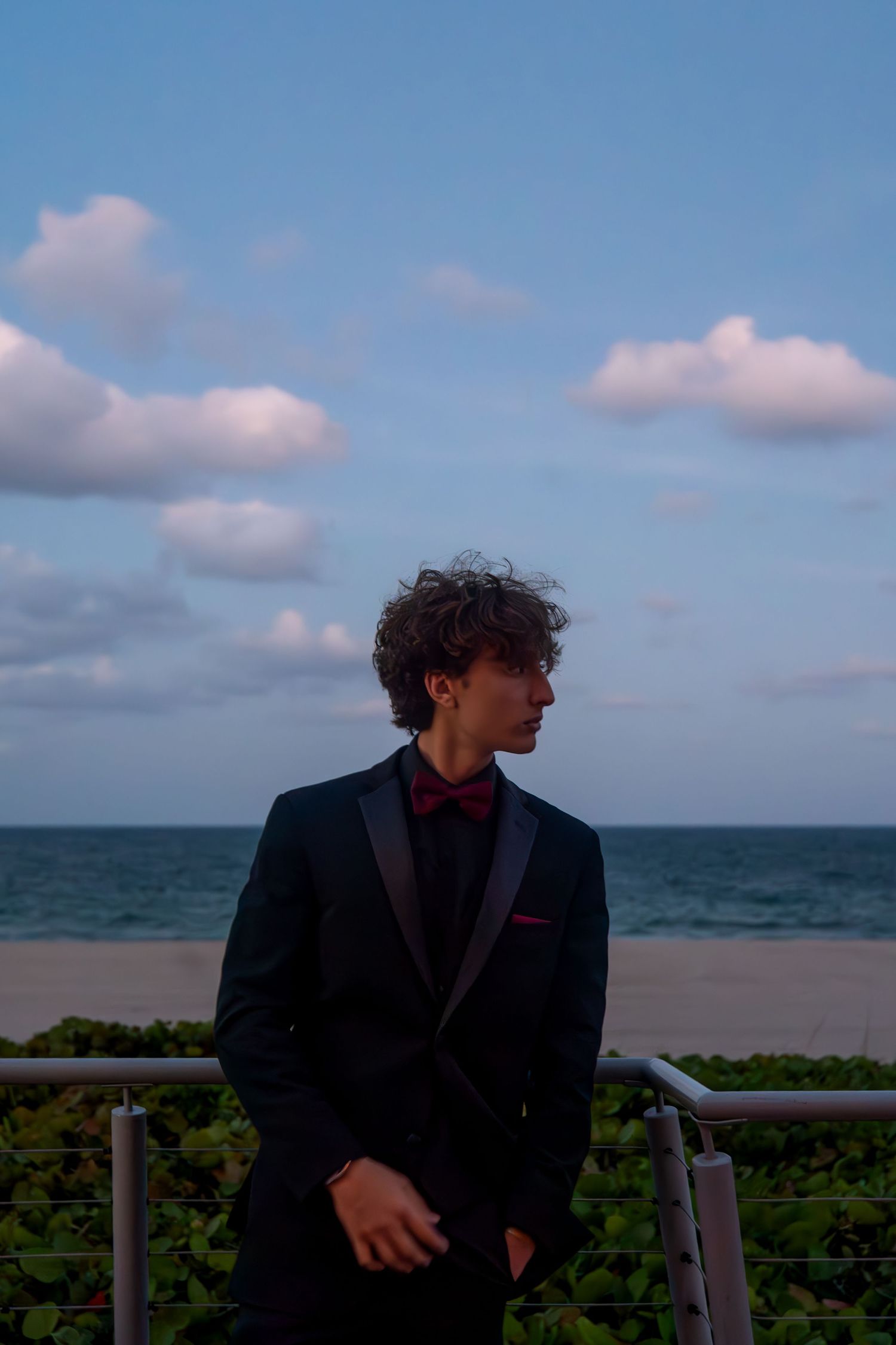 Person in dark suit stands alone against ocean backdrop with cloudy sky.