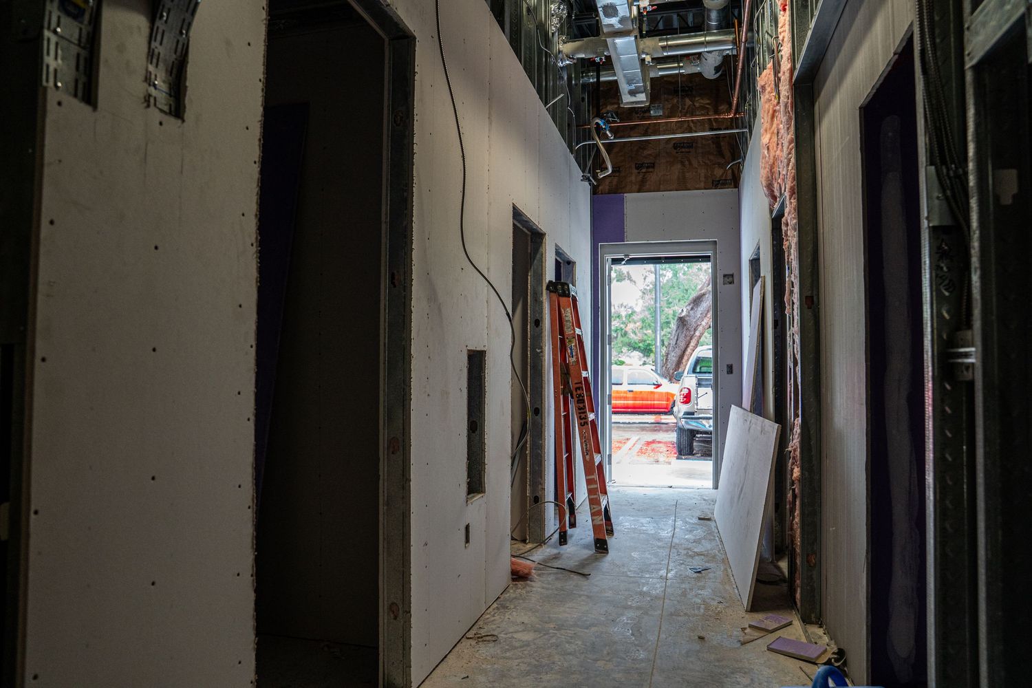 An under-construction hallway with exposed walls and ceiling infrastructure leads to a bright exit.