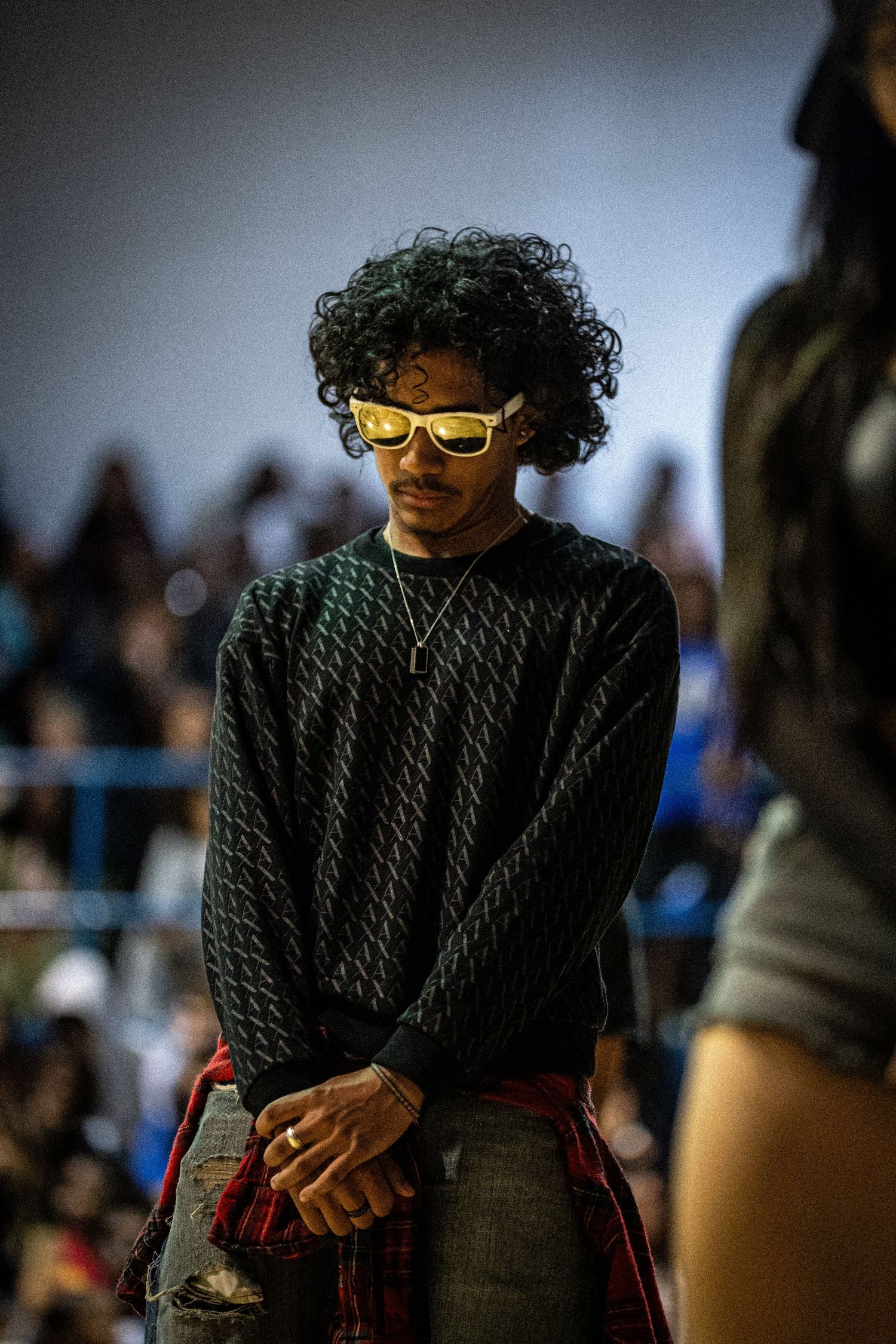 Young man with curly hair wearing yellow sunglasses and a dark patterned sweater at a fashion event.