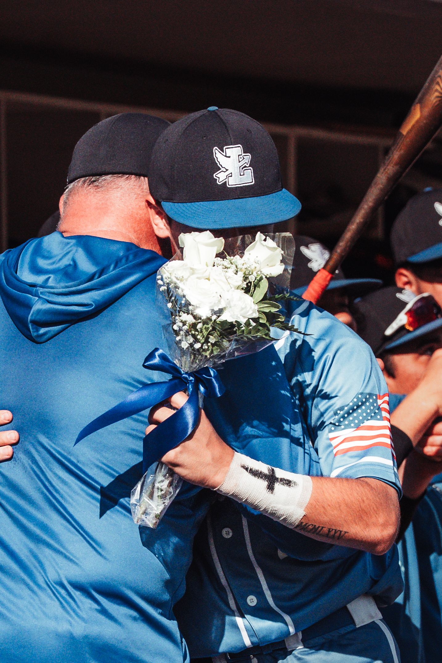 Baseball players in blue uniforms share an emotional hug, one holding a bouquet of white roses tied with a blue ribbon.