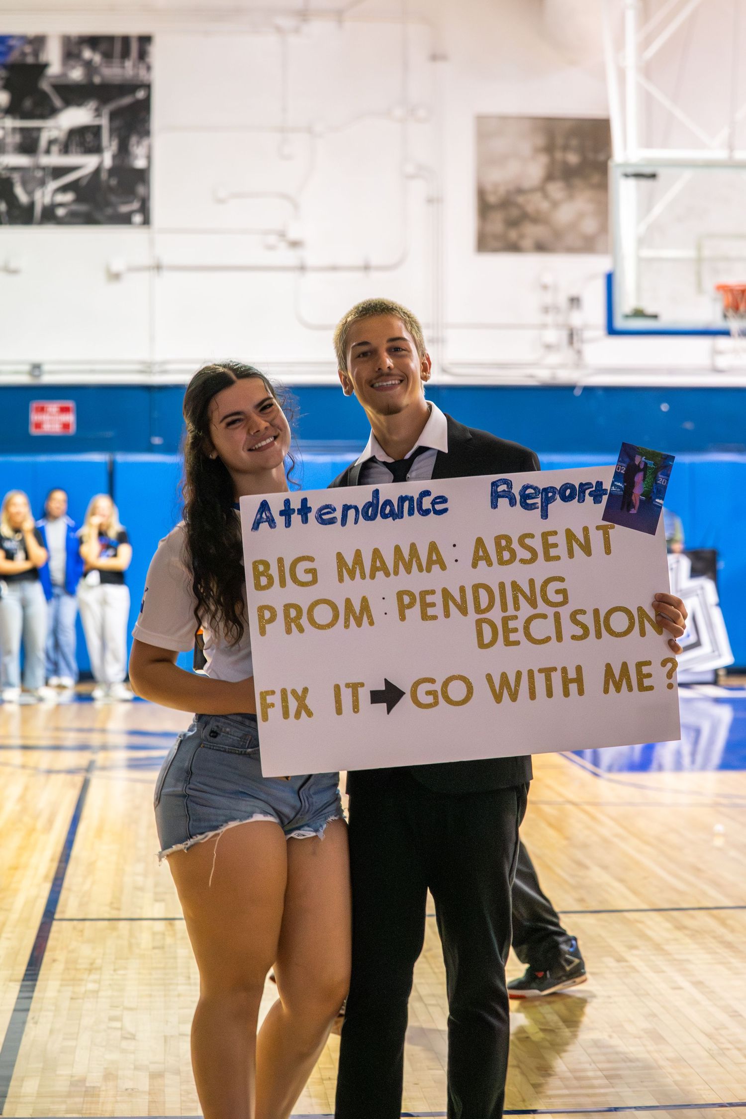 Two teens hold a creative promposal sign reading attendance report in a school gymnasium.
