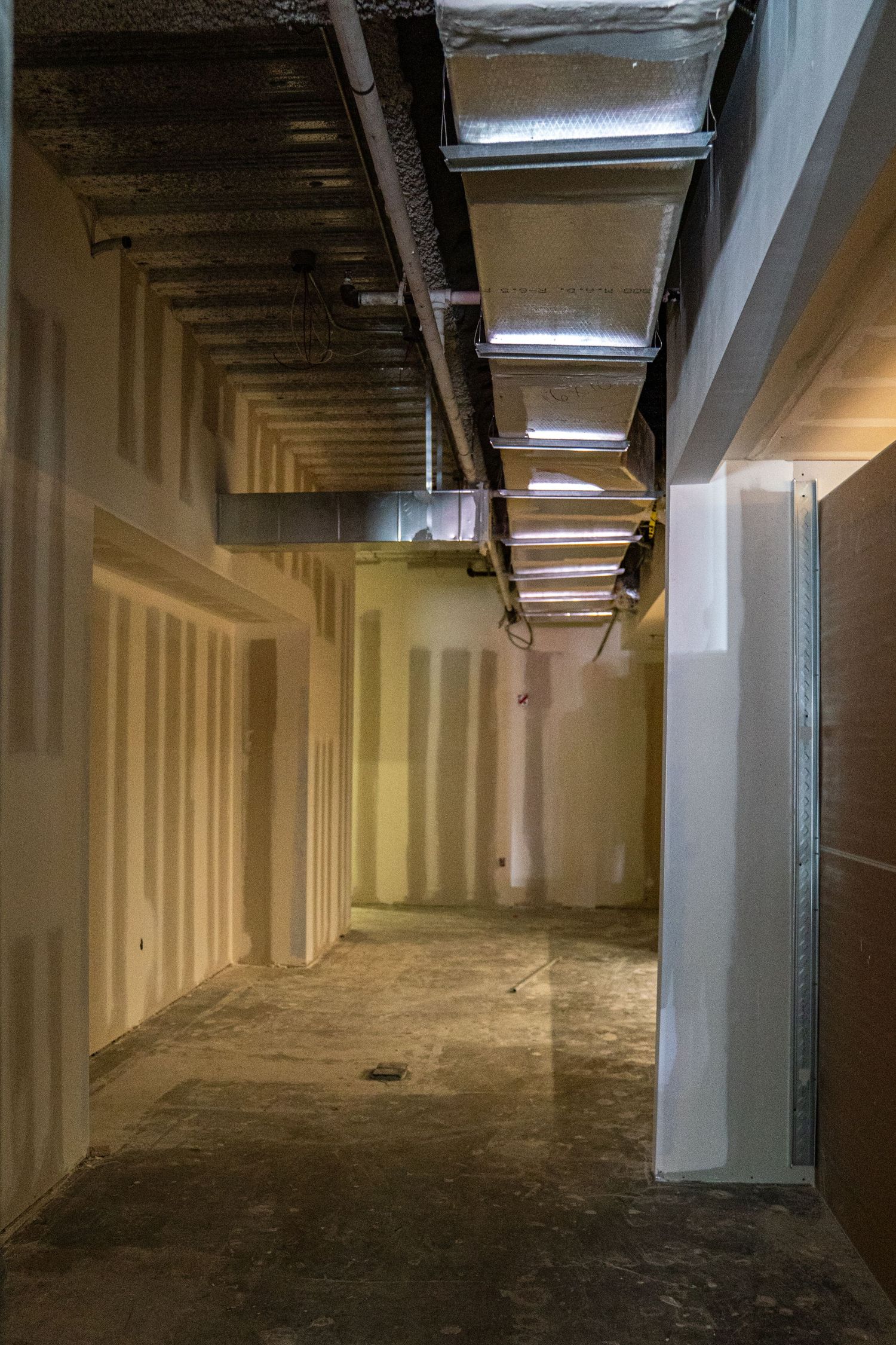 Dimly lit industrial corridor with exposed ceiling ducts and white walls.