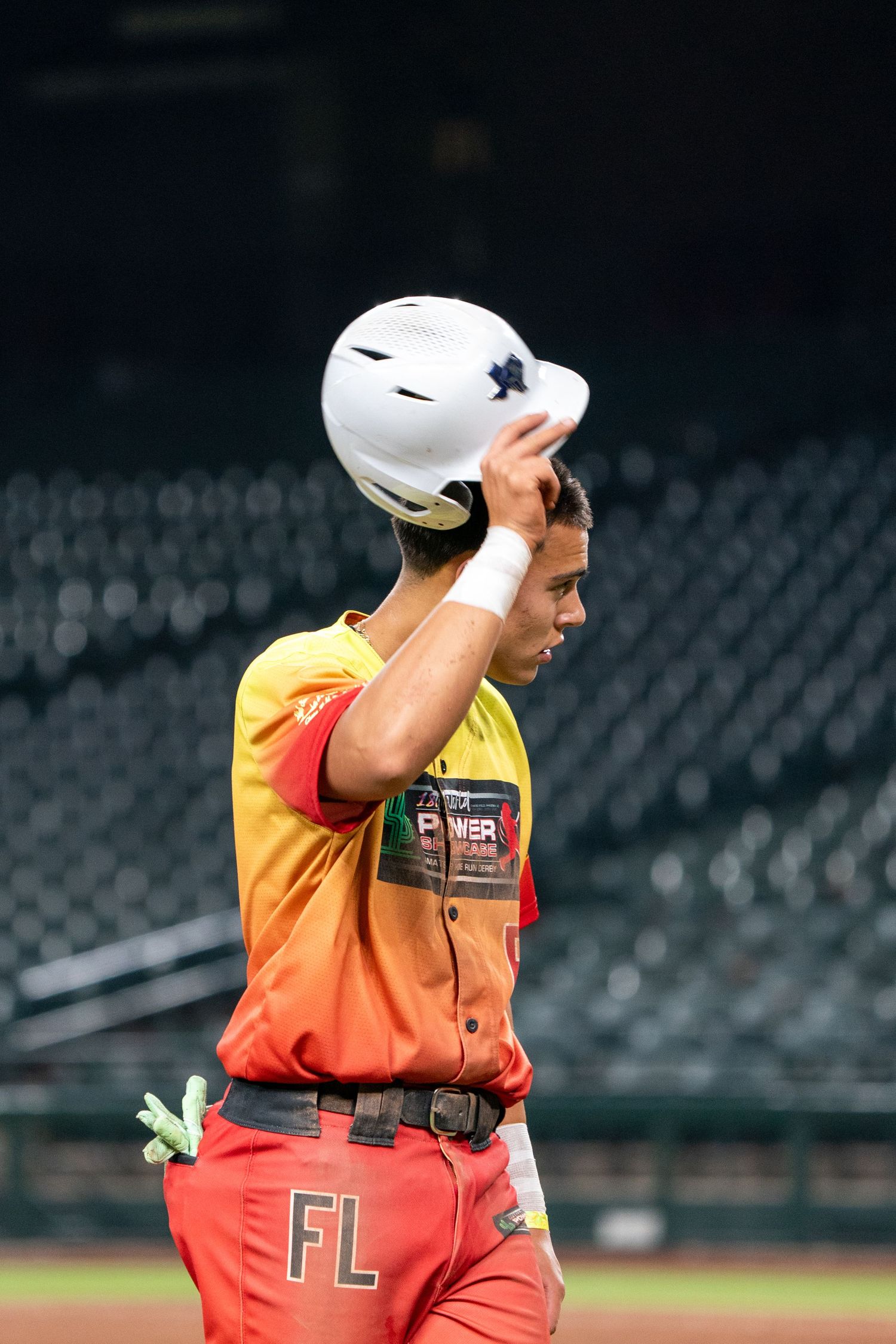 Baseball player in orange and red gradient uniform adjusts helmet while standing on field during game.