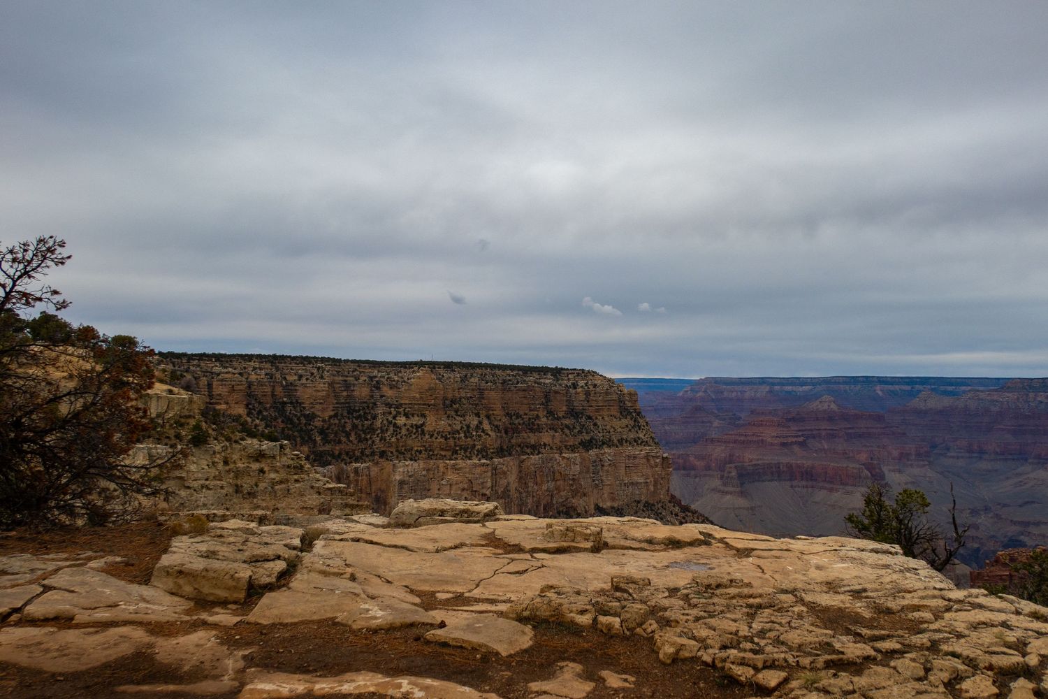 Dramatic overlook of the Grand Canyon showing vast cliffs and rock formations under an overcast sky.