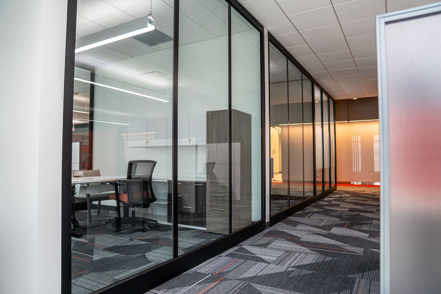 Corporate hallway with glass-enclosed offices, modern lighting, and geometric patterned flooring.