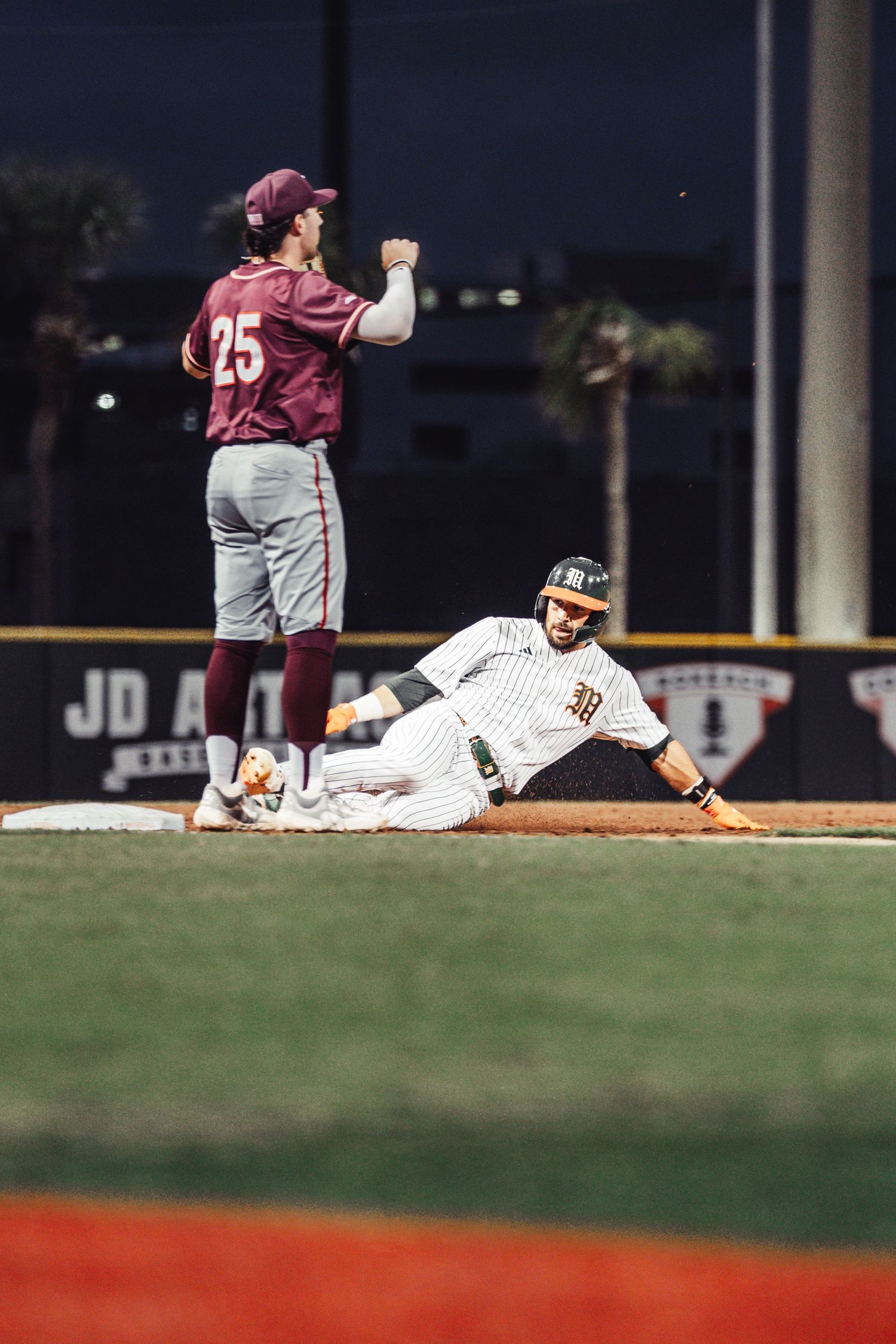 Baseball player slides into base as opponent in maroon jersey number 25 stands nearby during night game.