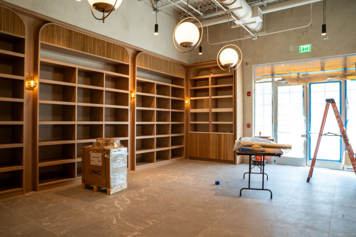 Empty wooden shelving units line the walls of an unfinished retail space with exposed ceiling and a ladder visible.