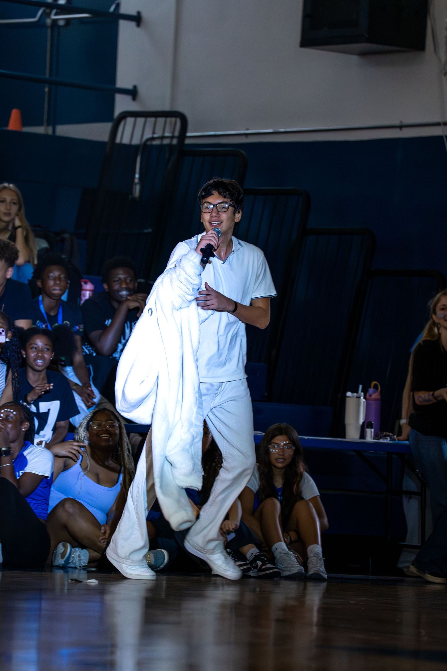 Young man in white outfit holds microphone performing at school pep rally in gymnasium with students seated in bleachers.