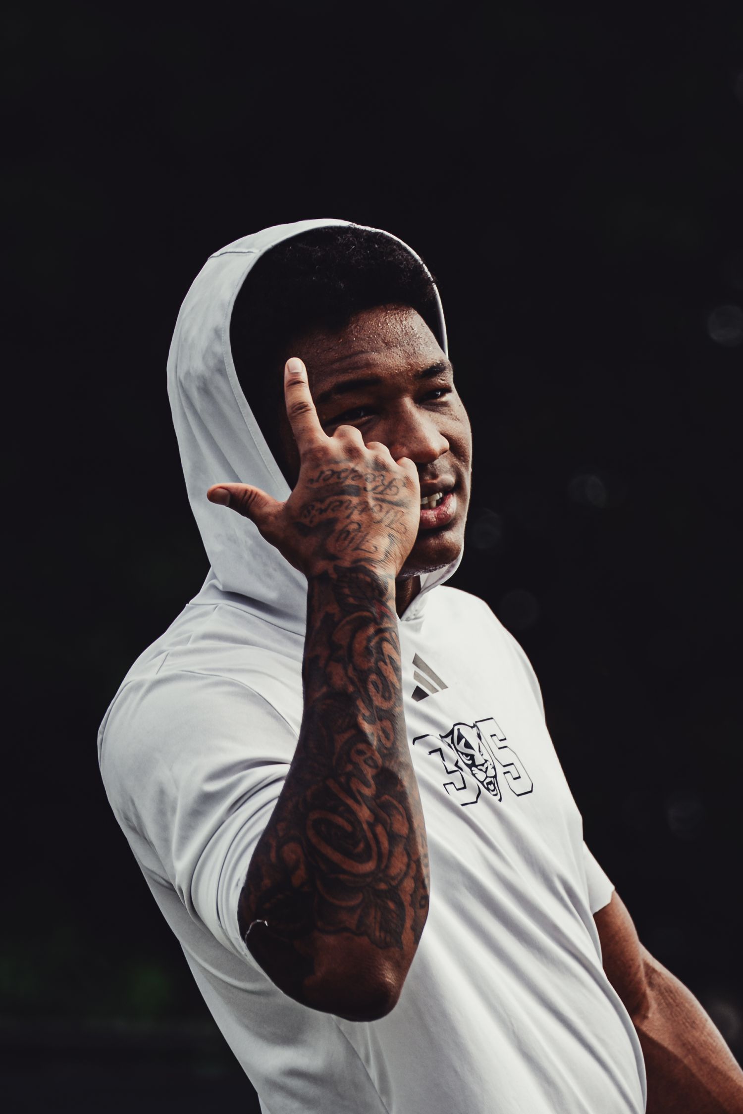 Man in white hoodie with tattooed arm making hand gesture against dark background.
