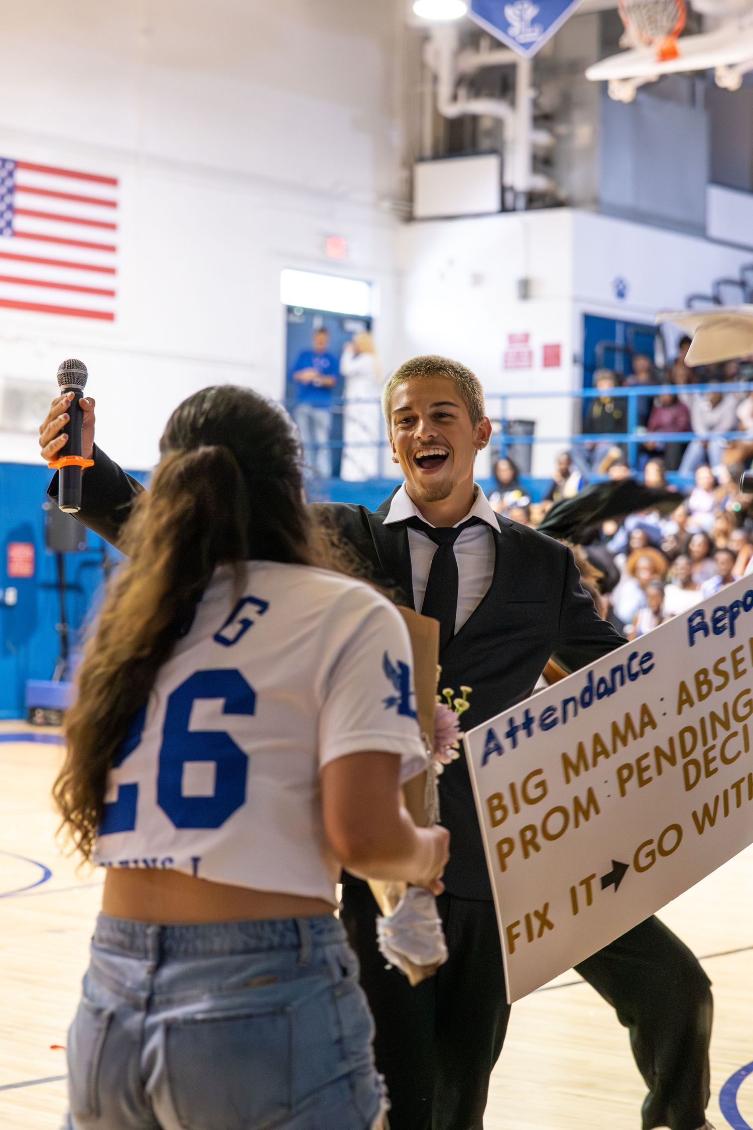 Student in suit holds mic laughing at prom proposal sign during high school gym pep rally event.