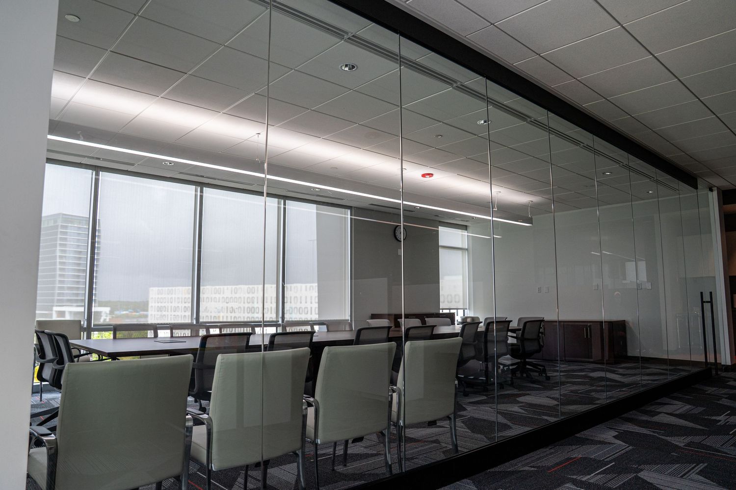 Minimalist conference room with glass walls, gray chairs, and linear ceiling lighting.