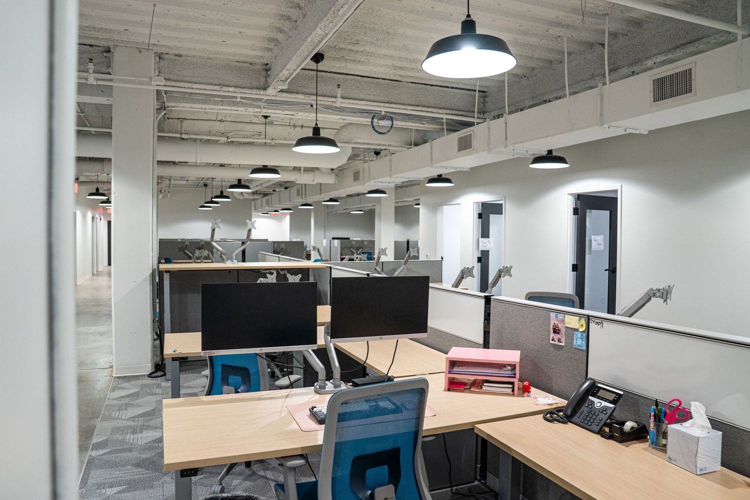 Modern open office space with exposed ceiling, pendant lighting, and light wood desks in a minimalist industrial setting.