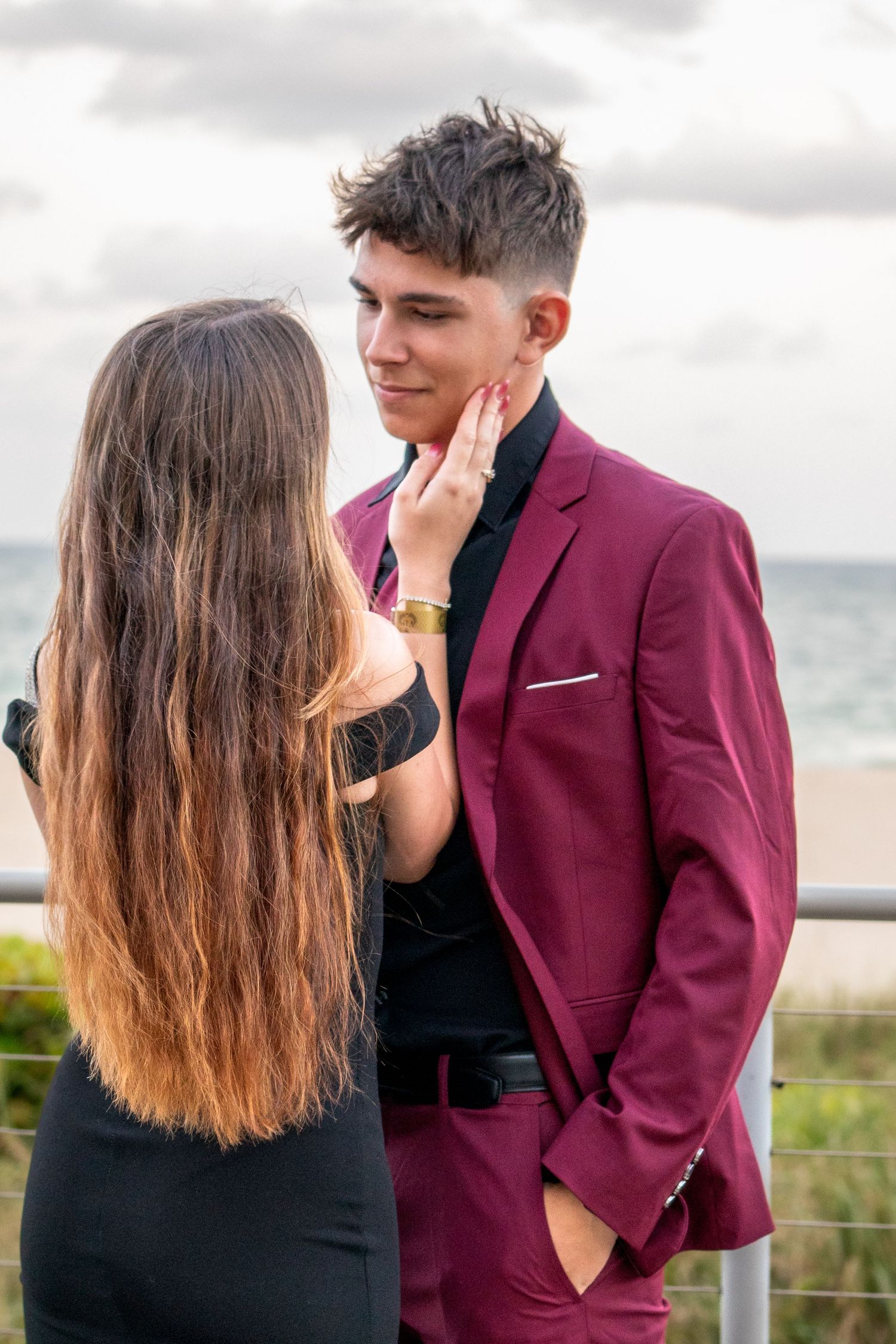 Couple shares intimate moment near beach, one wearing burgundy suit jacket.