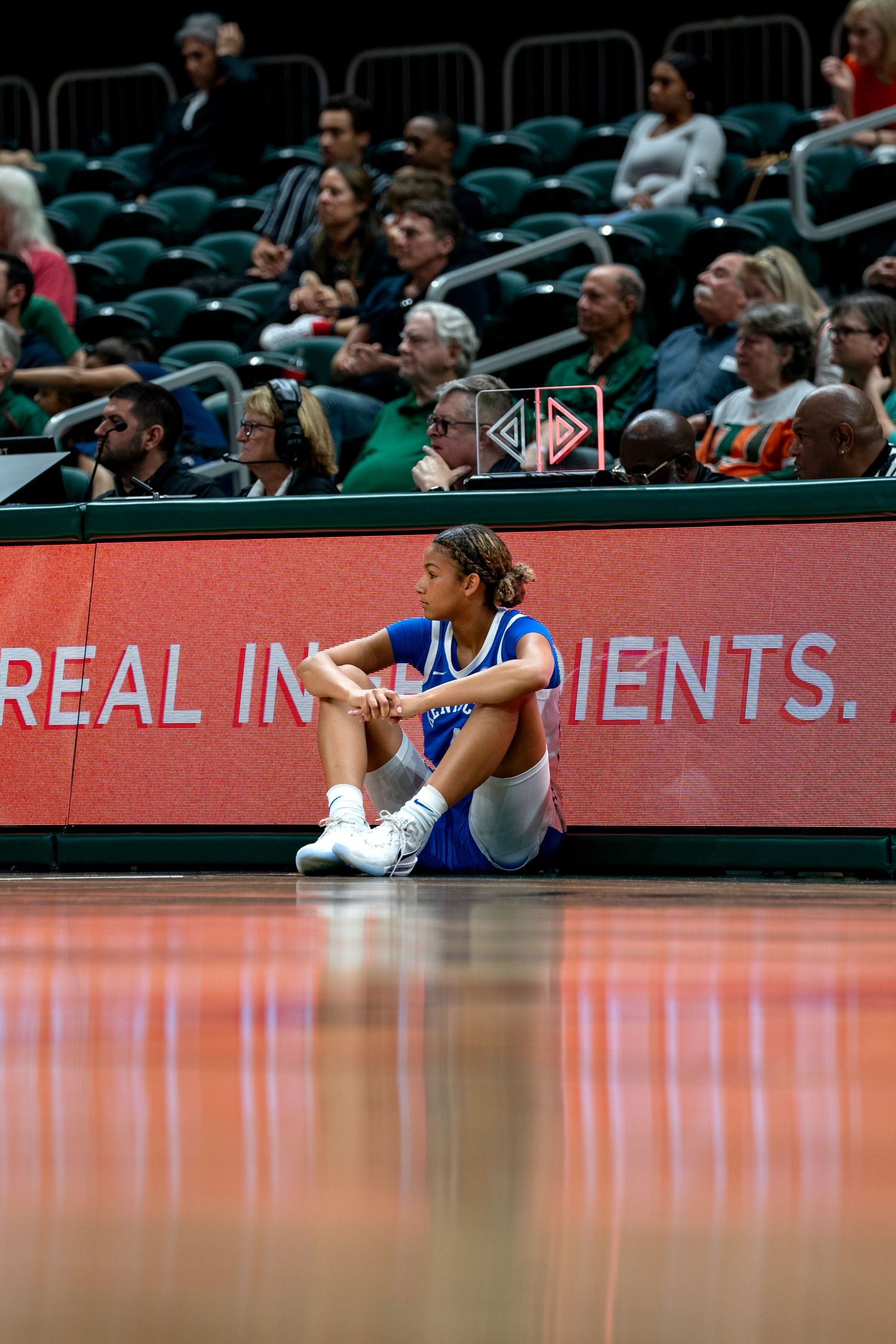 Basketball player sits on court near advertising board during game.