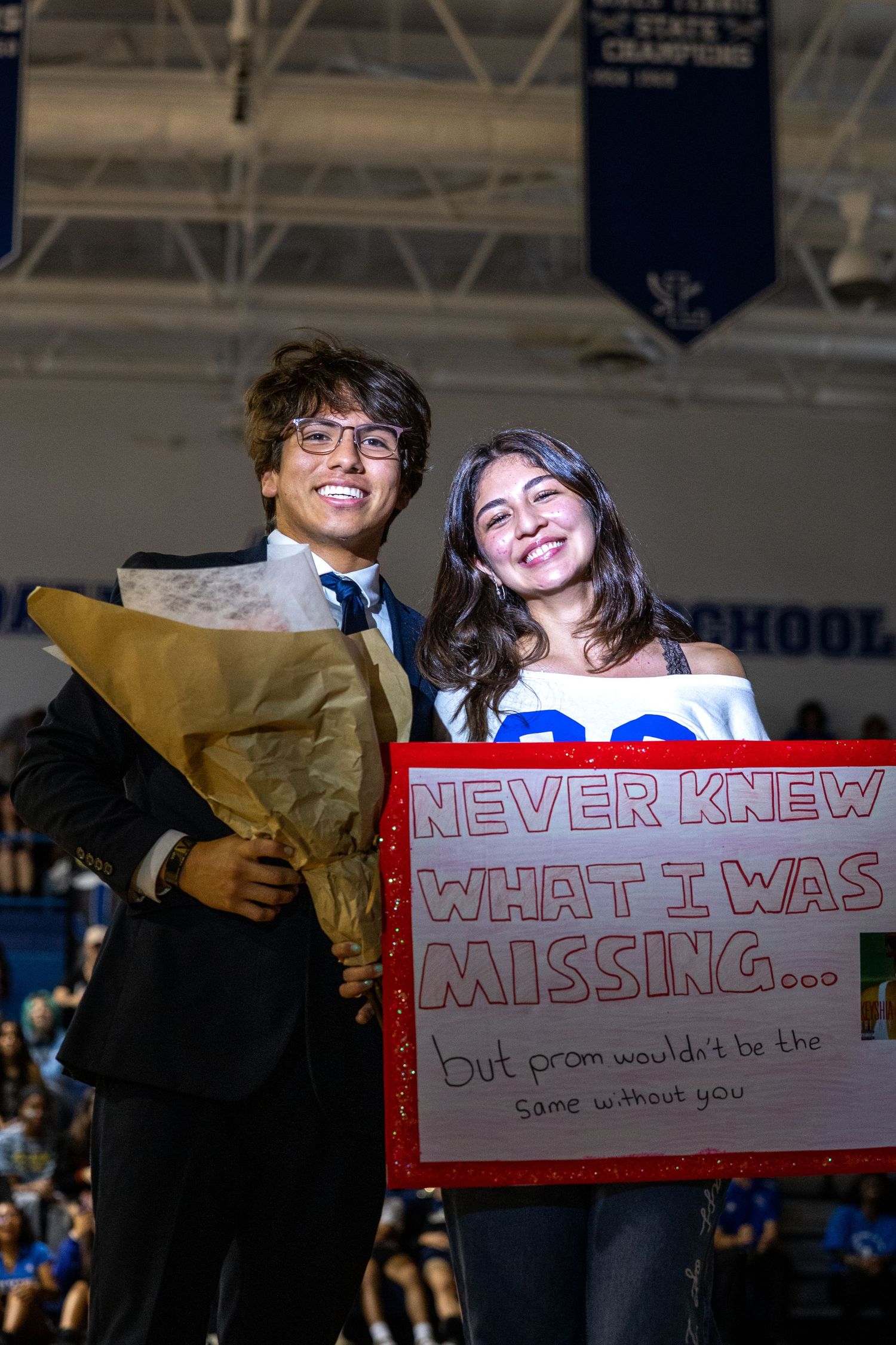 Two smiling high school students pose with a colorful promposal sign and flowers at a school event.