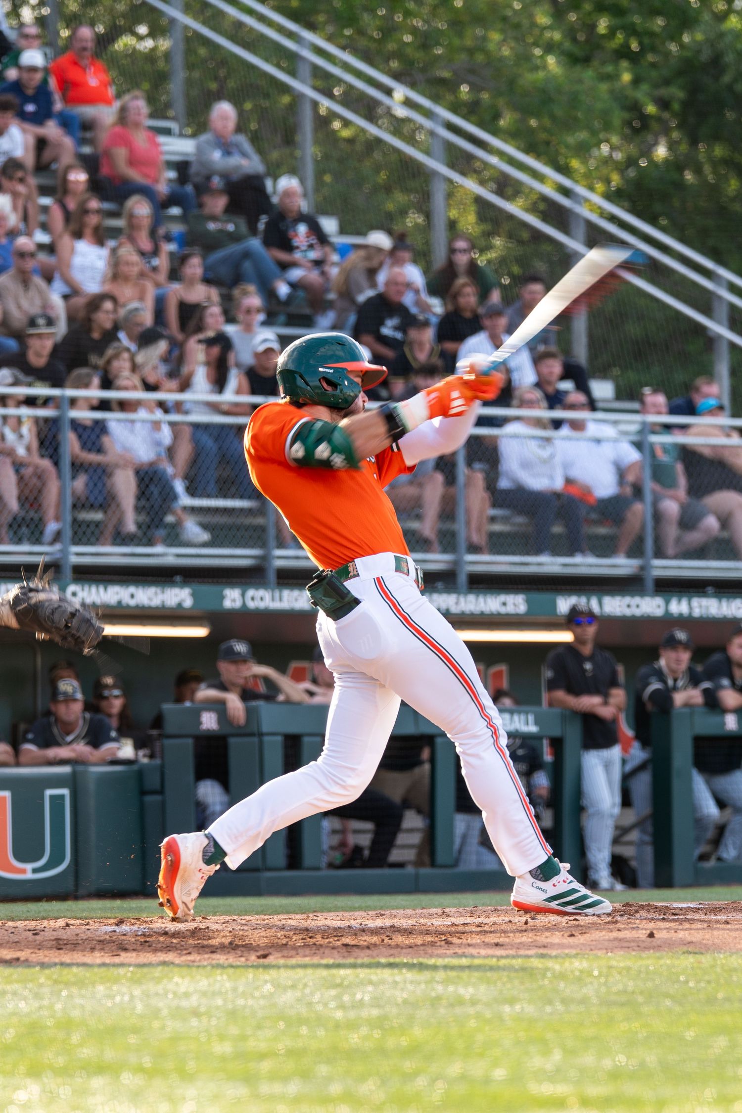 University of Miami baseball player in orange uniform swinging bat during a game with fans in background.