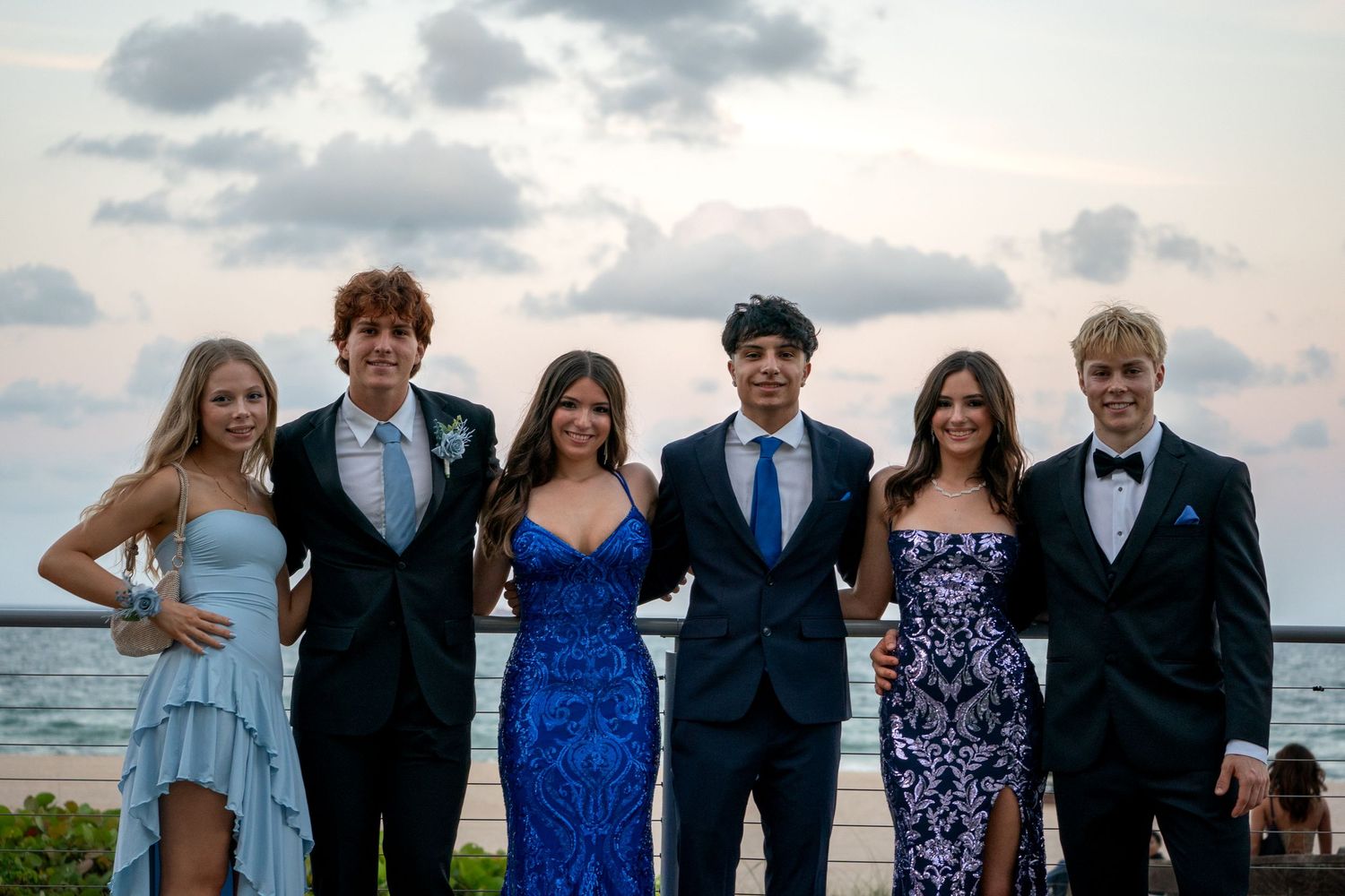 Group photo of formal event attendees posing together by the ocean at sunset.