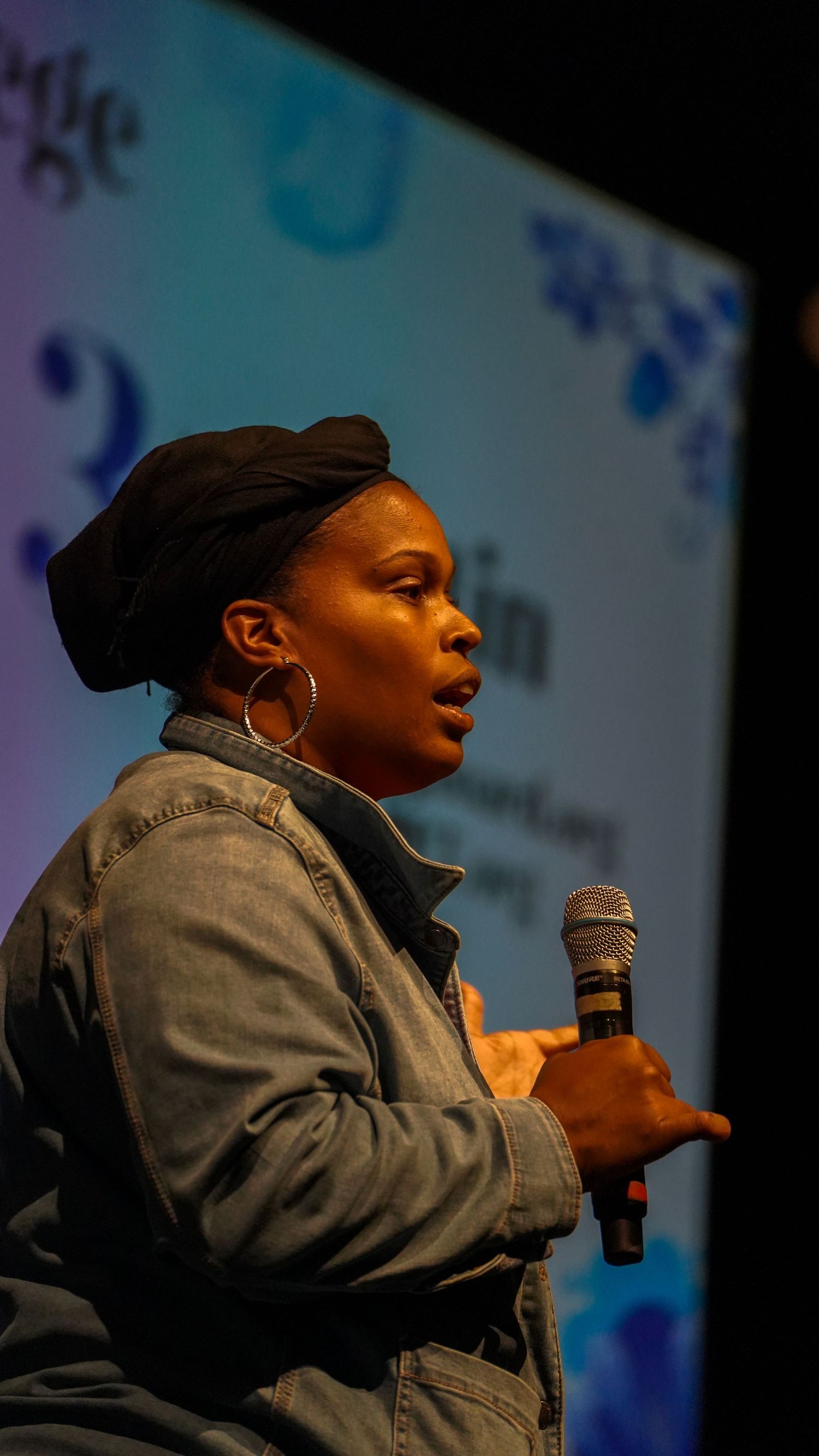 Speaker wearing denim jacket and hoop earrings presents against colorful backdrop.