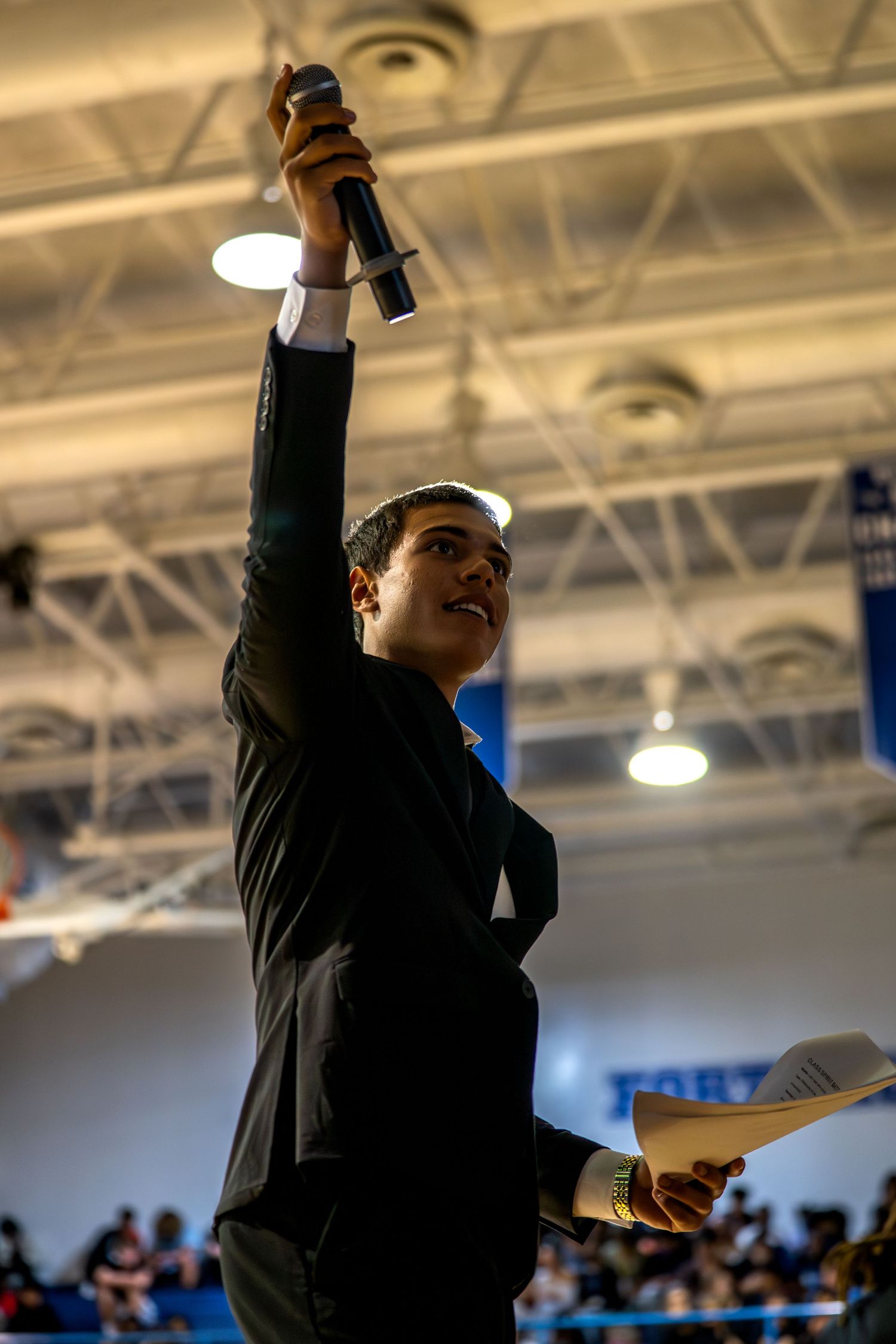 Person in black suit holds microphone high while speaking at an indoor arena event, holding papers in other hand.