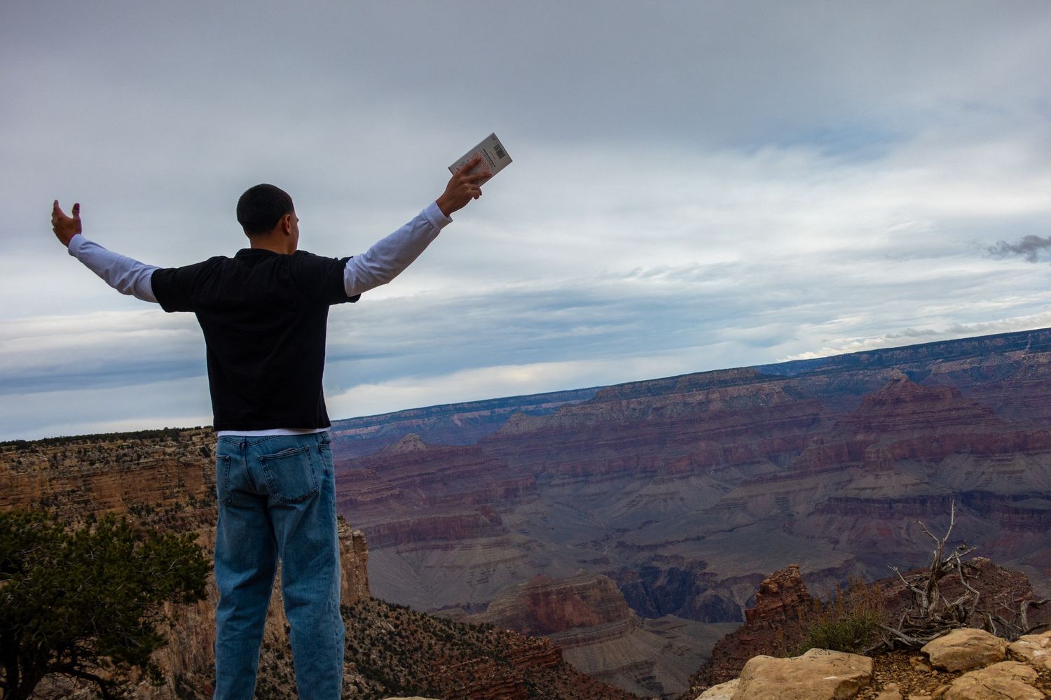 Person stands at the edge of the Grand Canyon with arms outstretched against a cloudy sky.