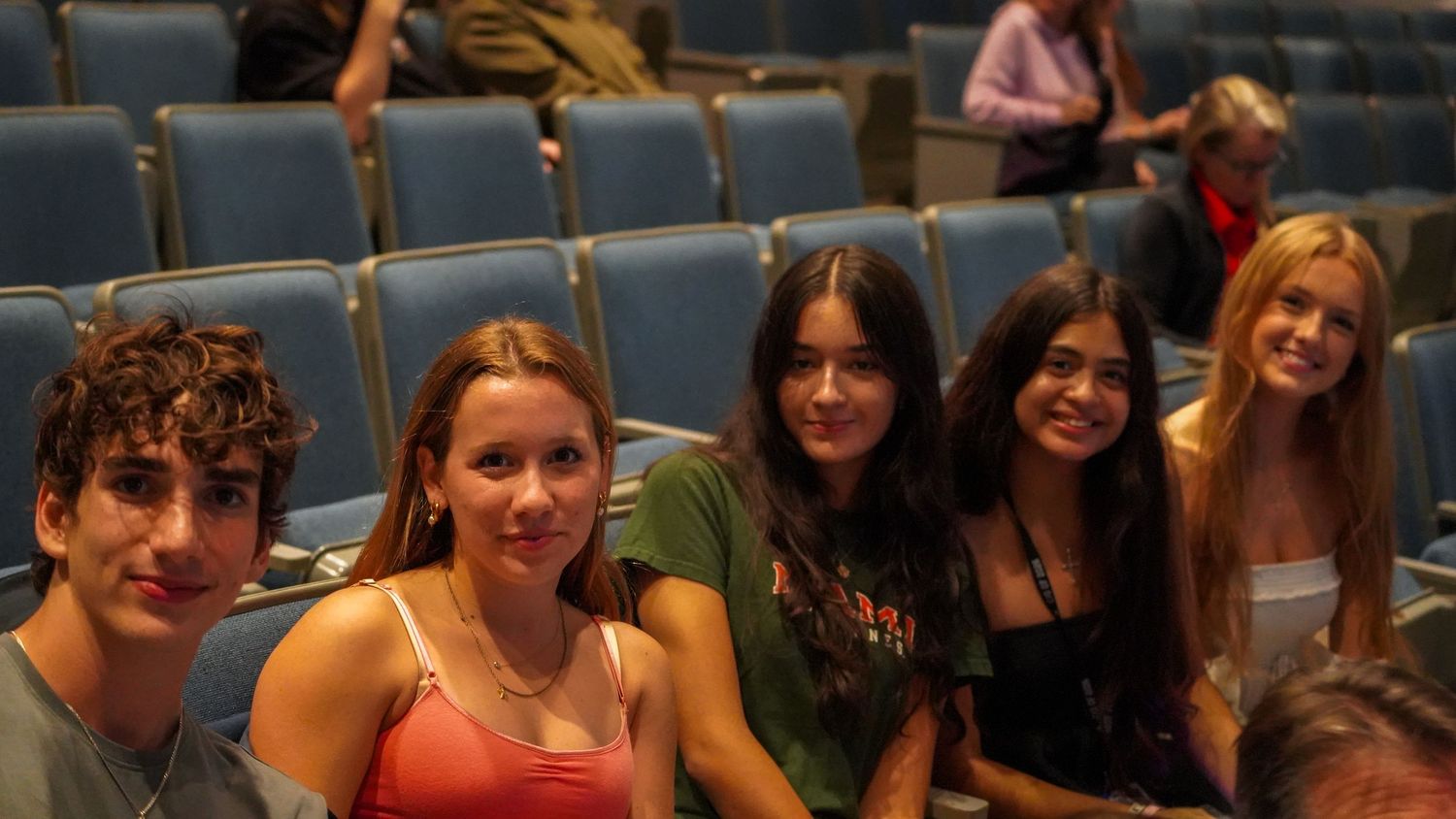 Students smiling and sitting together in theater-style seating during a school event.