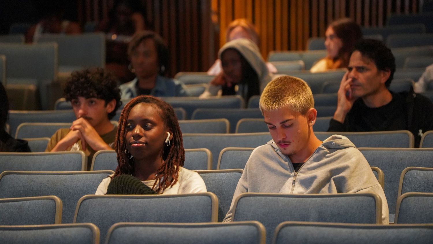 People seated in blue auditorium chairs watching a performance.