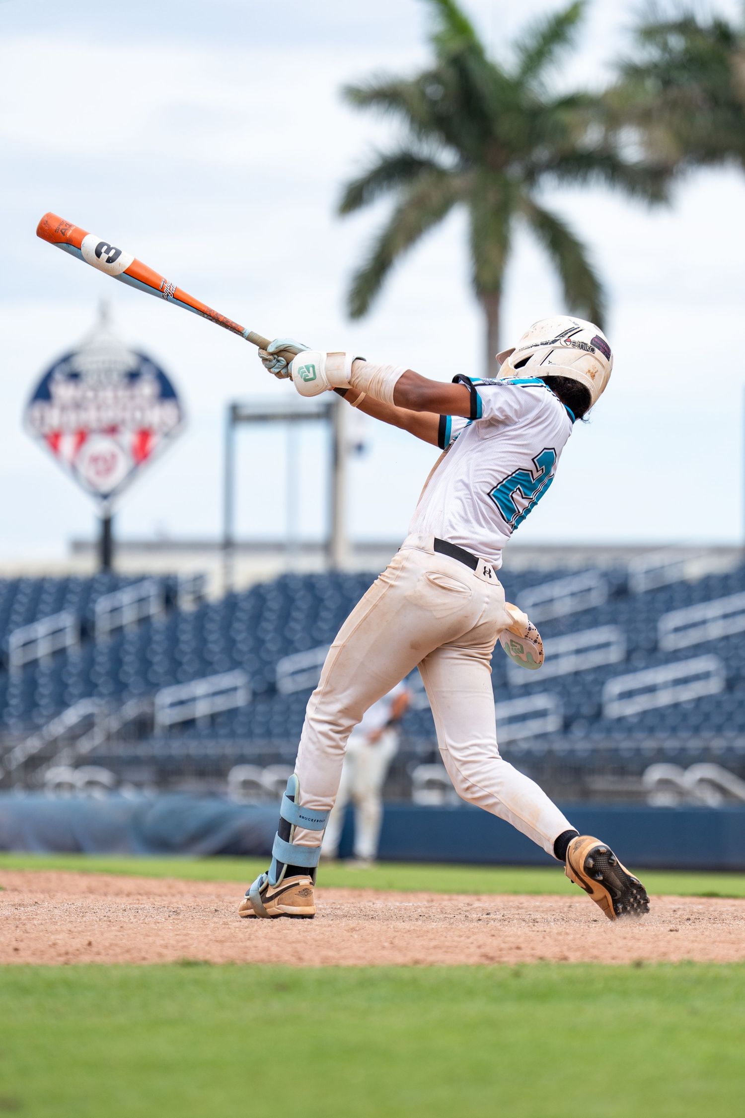Young baseball player in white uniform swings bat during a game at a professional stadium with palm trees.