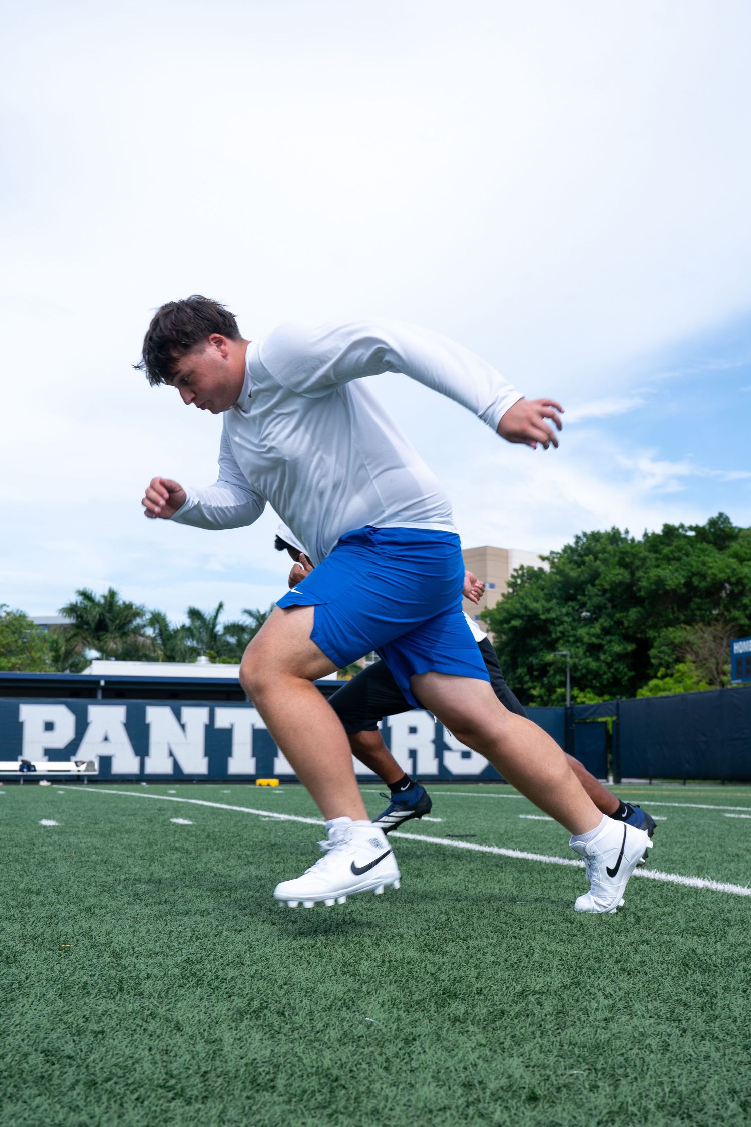 A football player in white jersey and blue shorts sprints on a turf field in front of a Panthers sign.