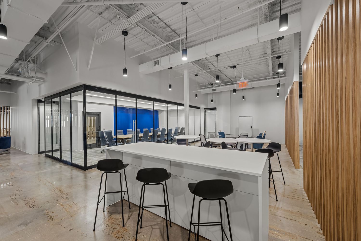 Modern open office space with white walls, exposed ceiling, wood slat dividers and black bar stools along counter seating.