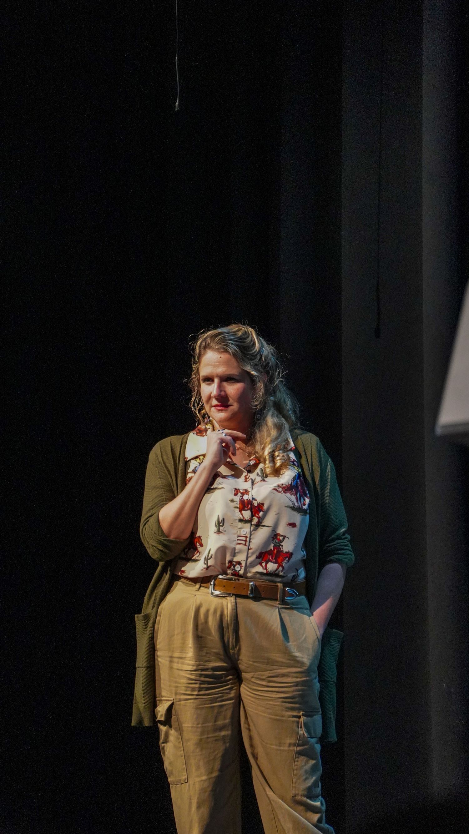 A performer in khaki pants and floral top stands on stage in dramatic lighting.
