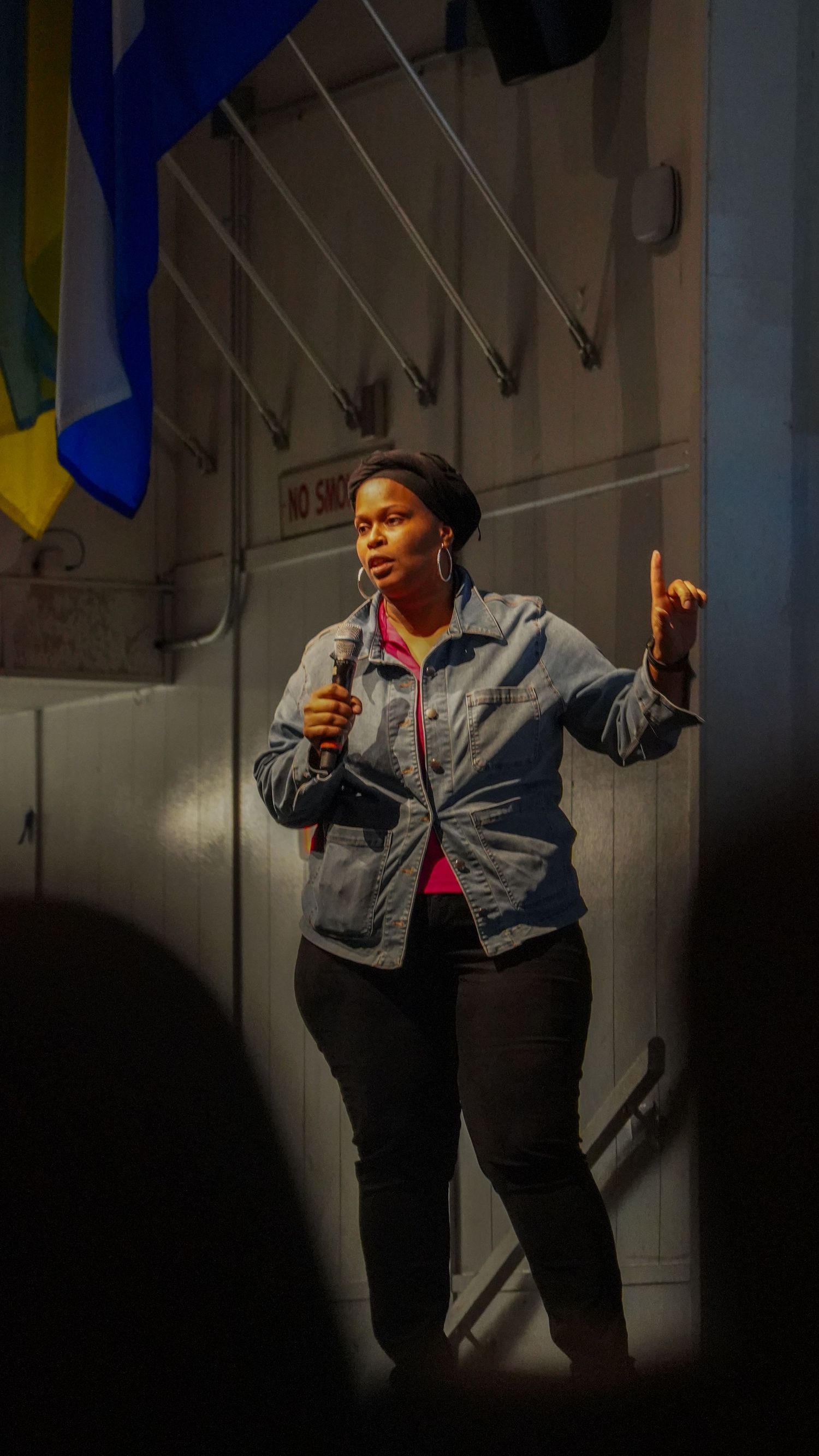 A performer in denim jacket gestures while speaking into microphone on stage.