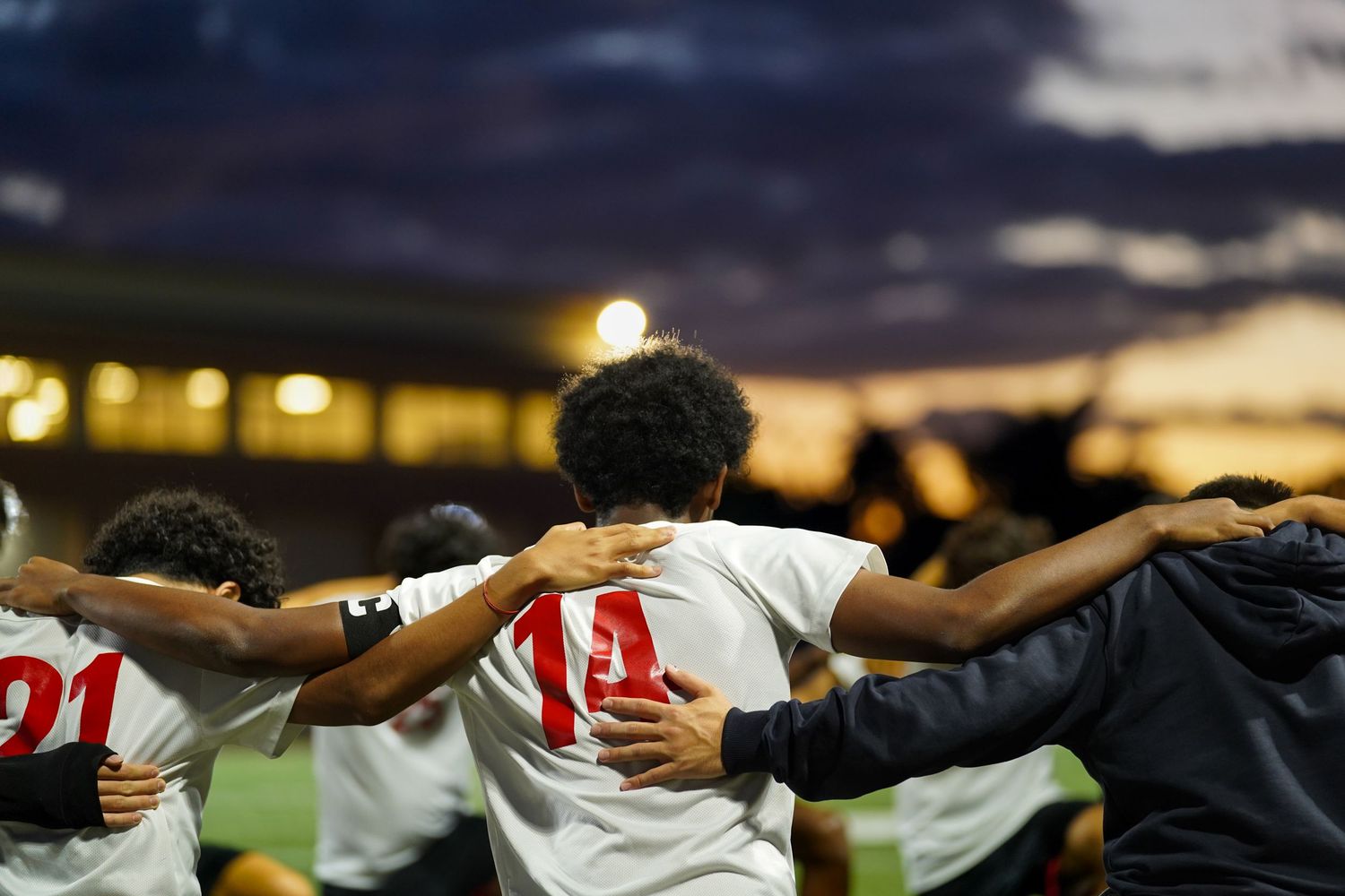 Soccer team stands with arms linked in solidarity during evening game.