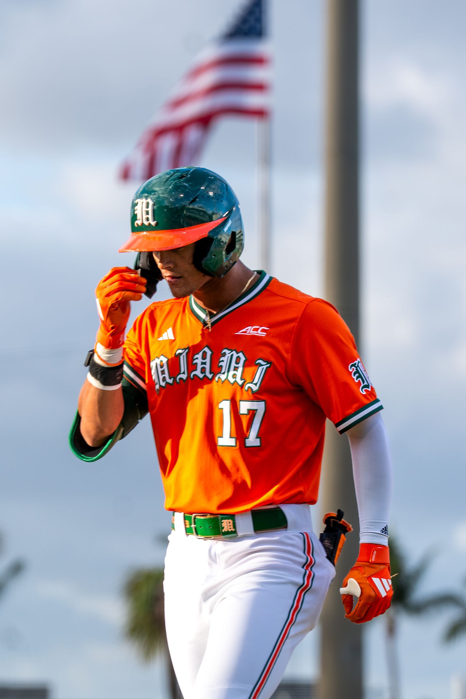 Miami Hurricanes baseball player #17 in orange uniform adjusts helmet near American flag.