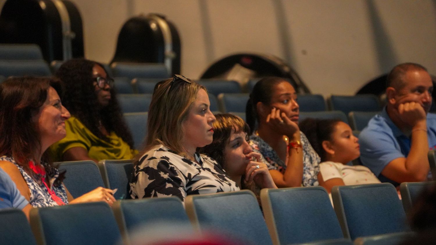 Audience members seated in blue chairs watch intently during a presentation.