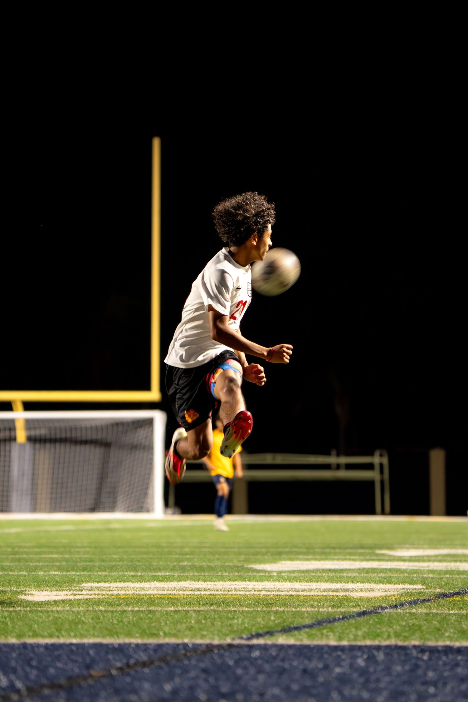 Soccer player leaps high to head the ball during nighttime match on artificial turf.