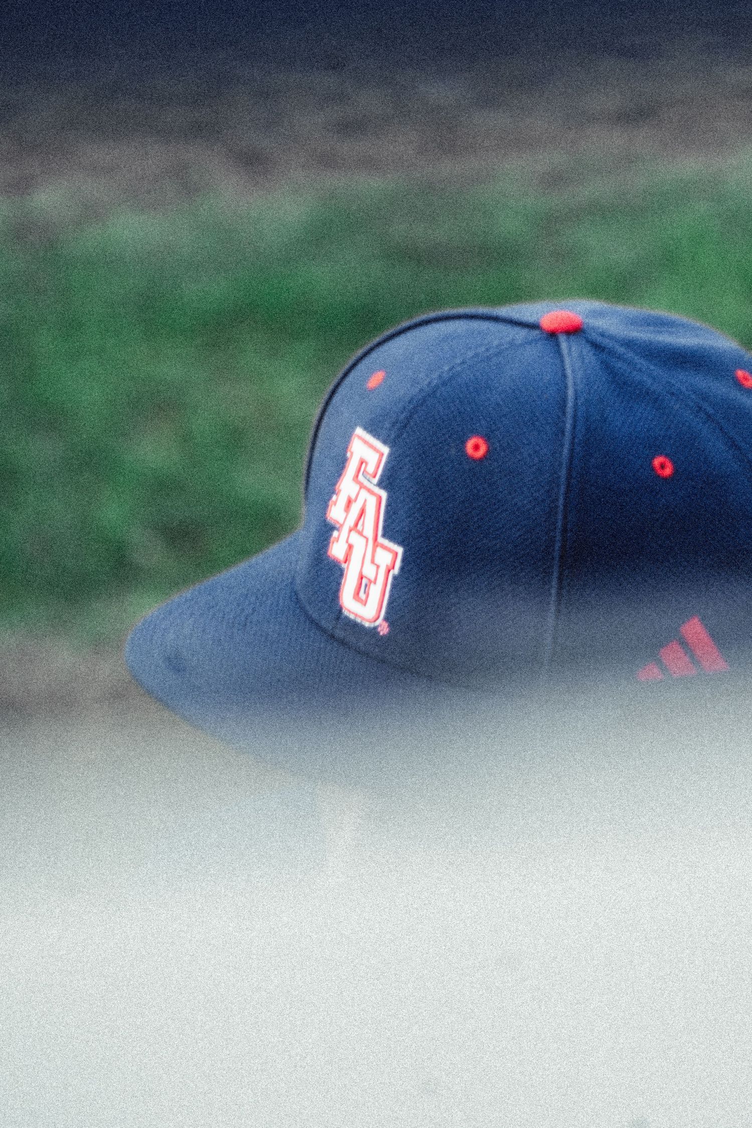 Navy blue FAU Adidas baseball cap with red logo resting on dugout surface.