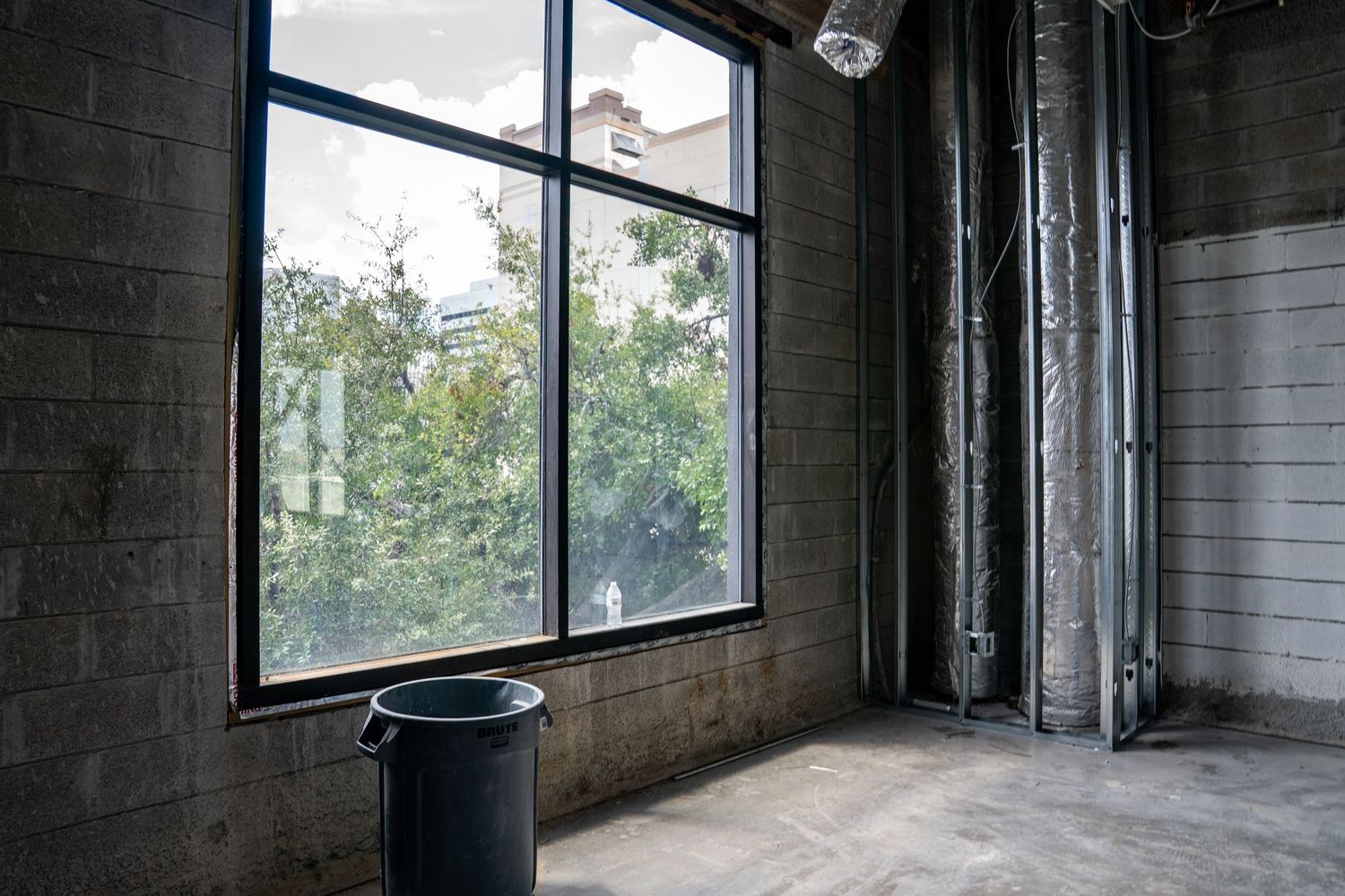 Urban view through large windows in an industrial space with exposed pipes and concrete walls.