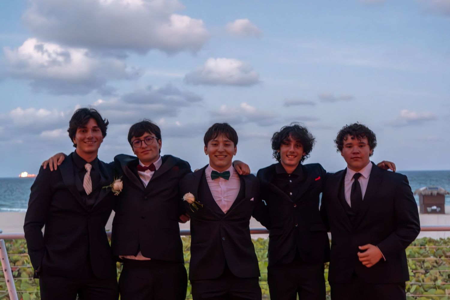 Group wearing formal attire poses together by the ocean on a cloudy day.