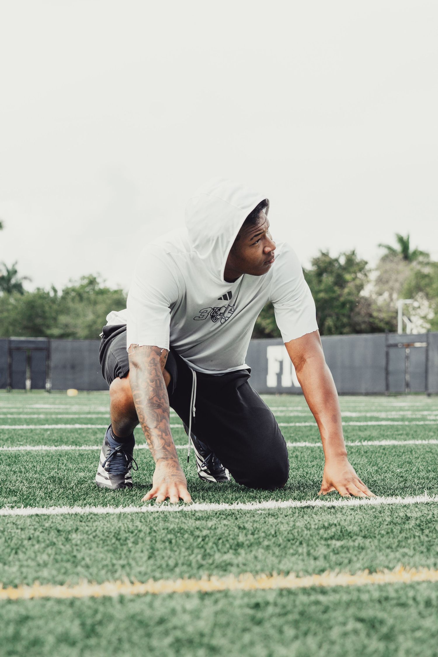 Athlete in Adidas hoodie crouches in starting position on green turf field, ready to sprint.