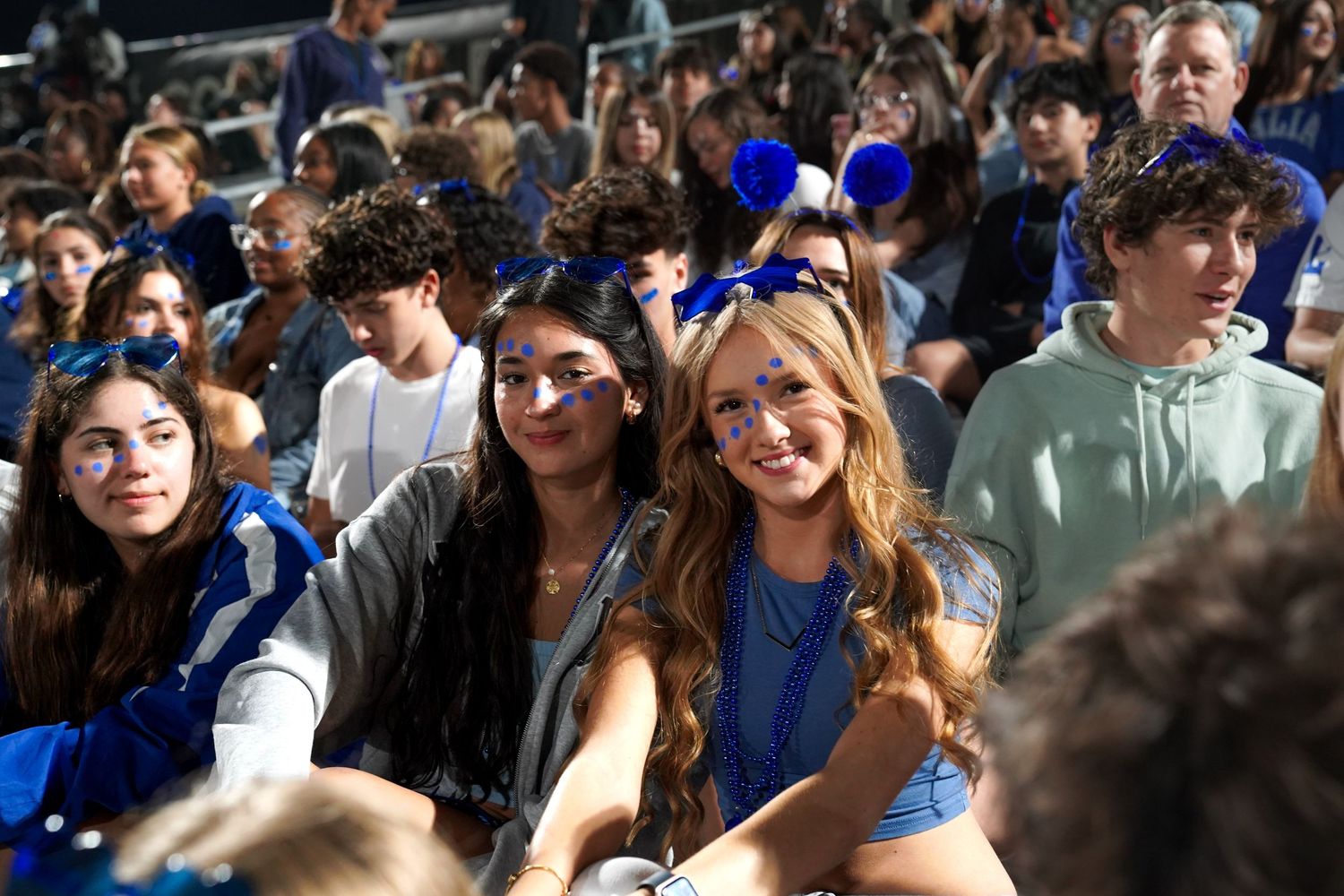Energetic student section wearing blue and white fills the stadium stands at night.
