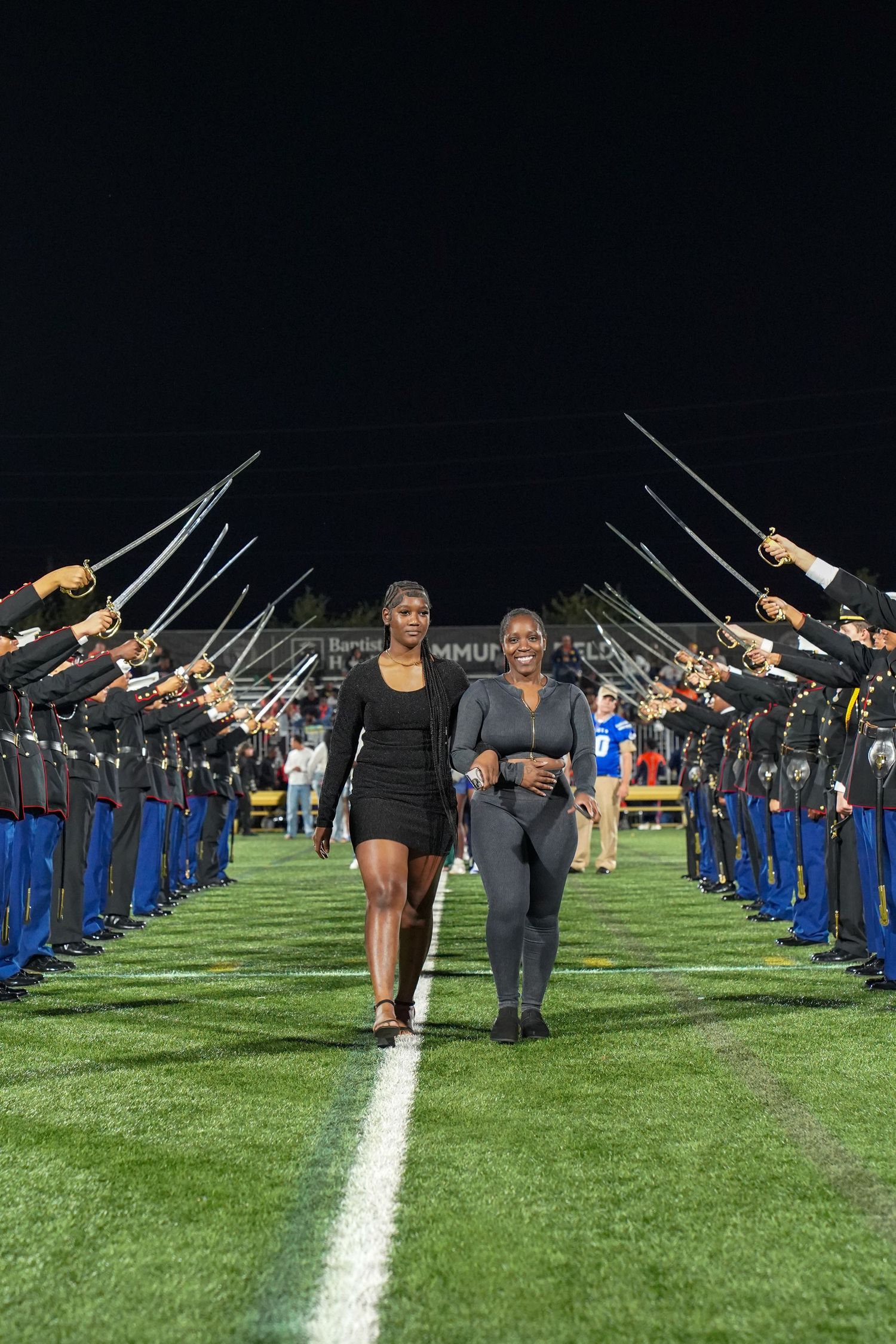 Students form two lines on a football field with raised props during a nighttime performance.