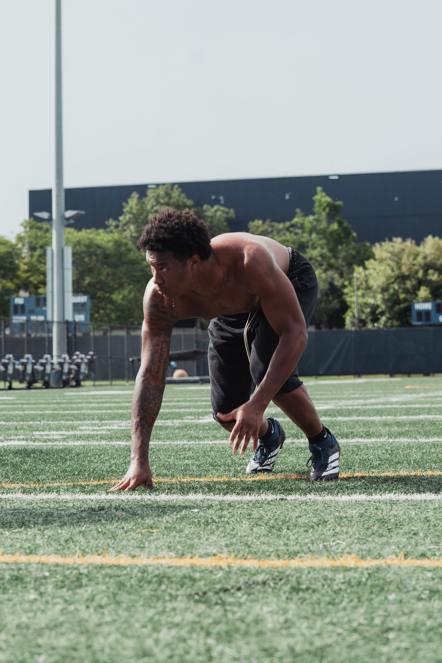Shirtless male athlete in a sprinter's starting stance on a green turf football field during outdoor training.