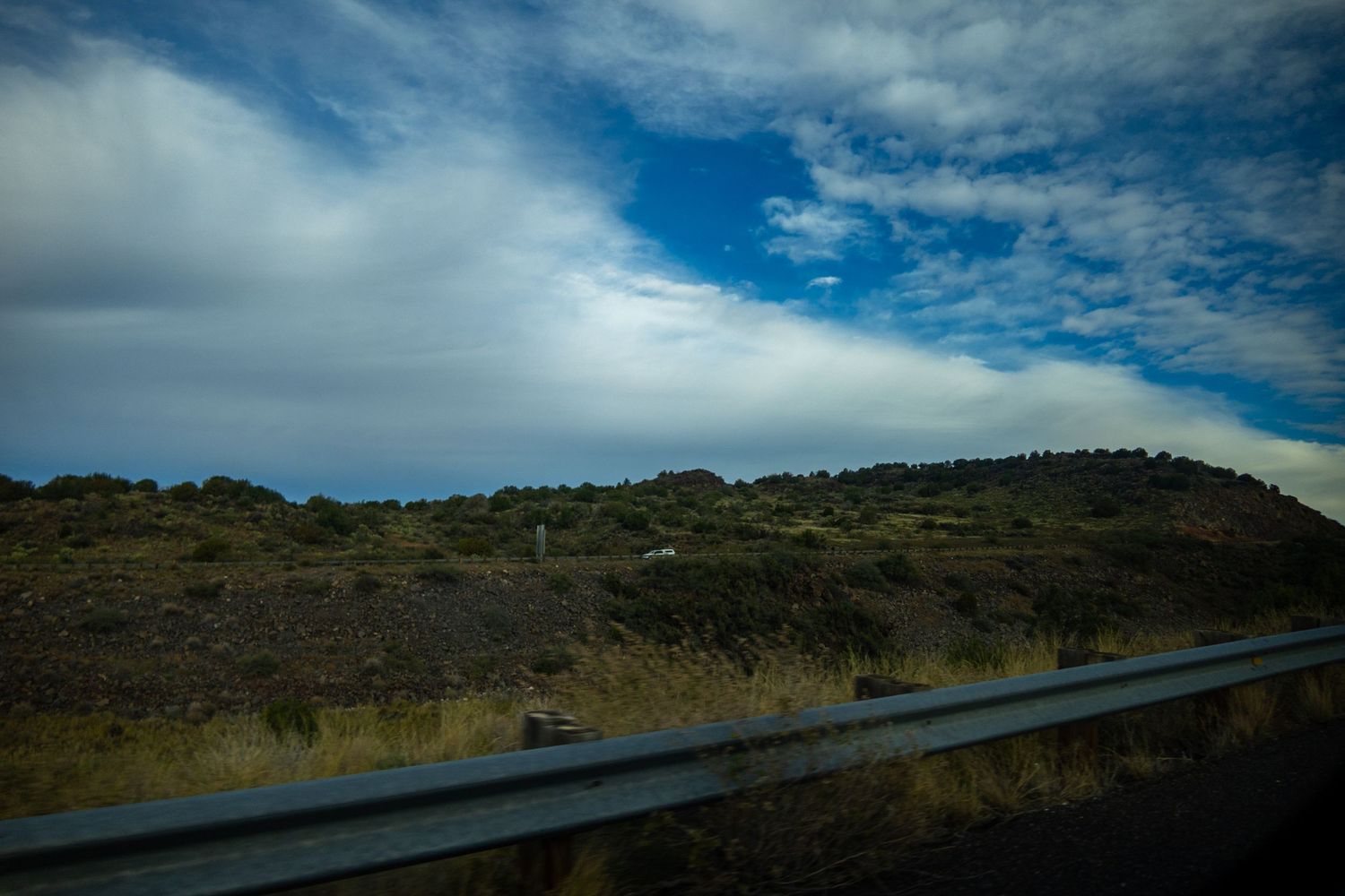 Desert road stretching across arid terrain with hills and sparse vegetation in the background.