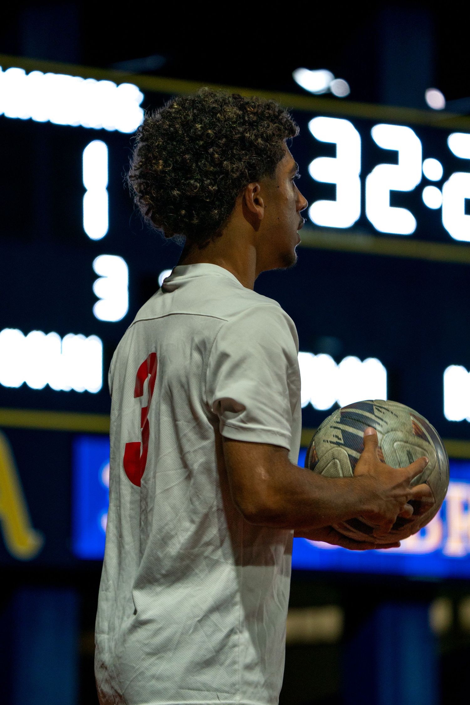 Basketball player holding ball during night game with scoreboard visible.