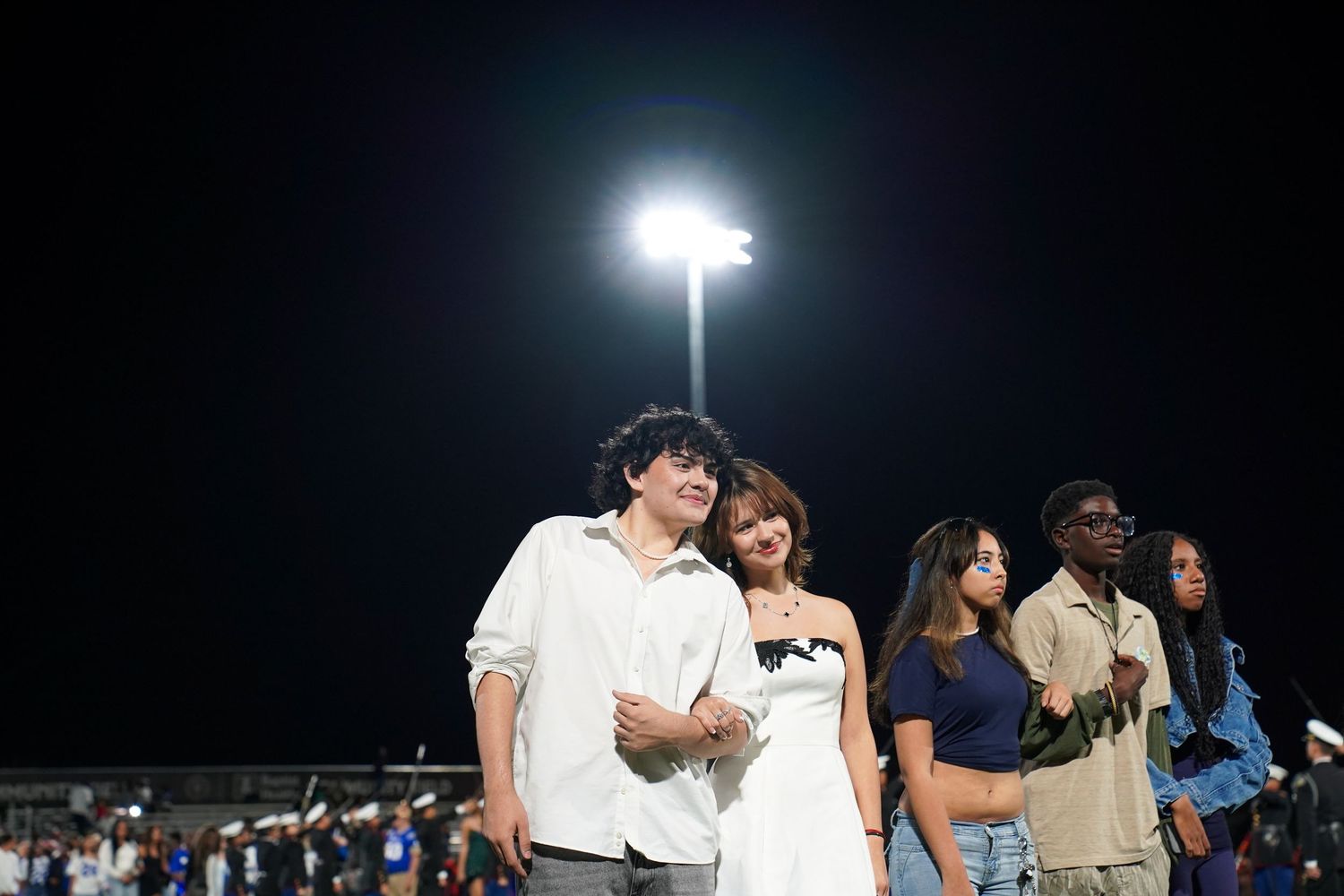 Students gather on a sports field at night under bright stadium lights for an outdoor event.