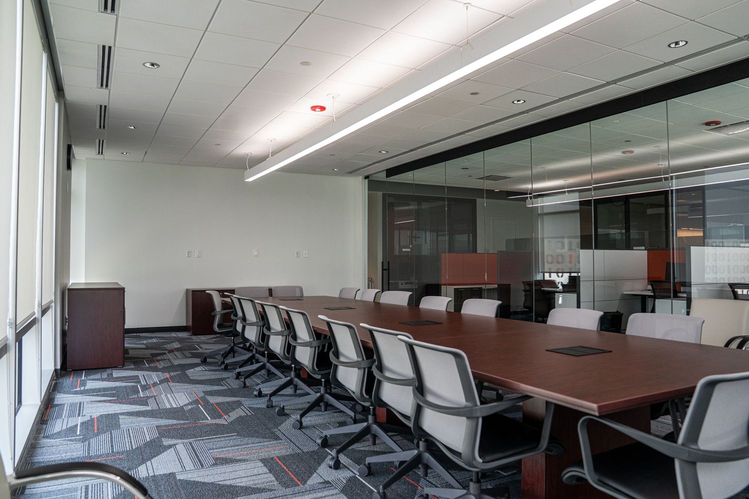 Modern conference room with long wooden table, ergonomic chairs, and glass walls featuring geometric patterned carpet.
