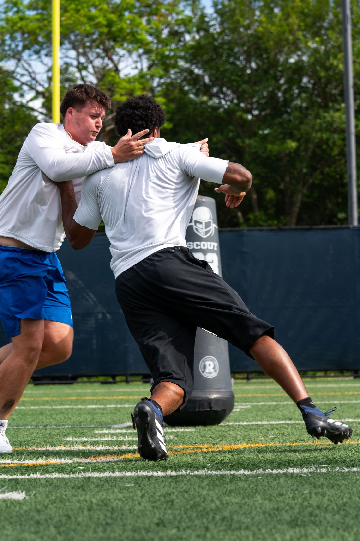 Two football players in white jerseys practice blocking drills against a tackle dummy on a green turf field.