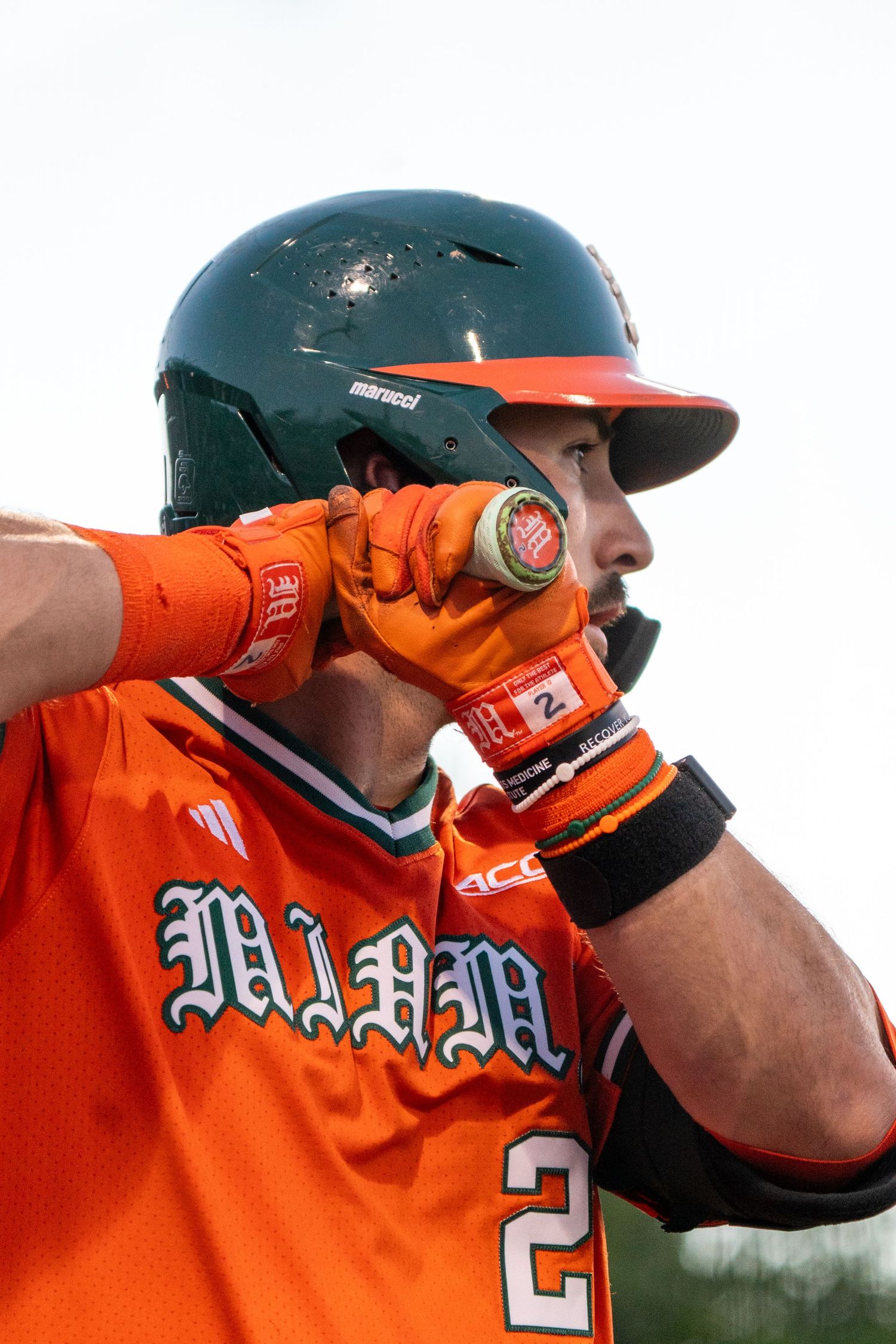 A Miami Hurricanes baseball player in an orange uniform adjusts his green helmet during a game.