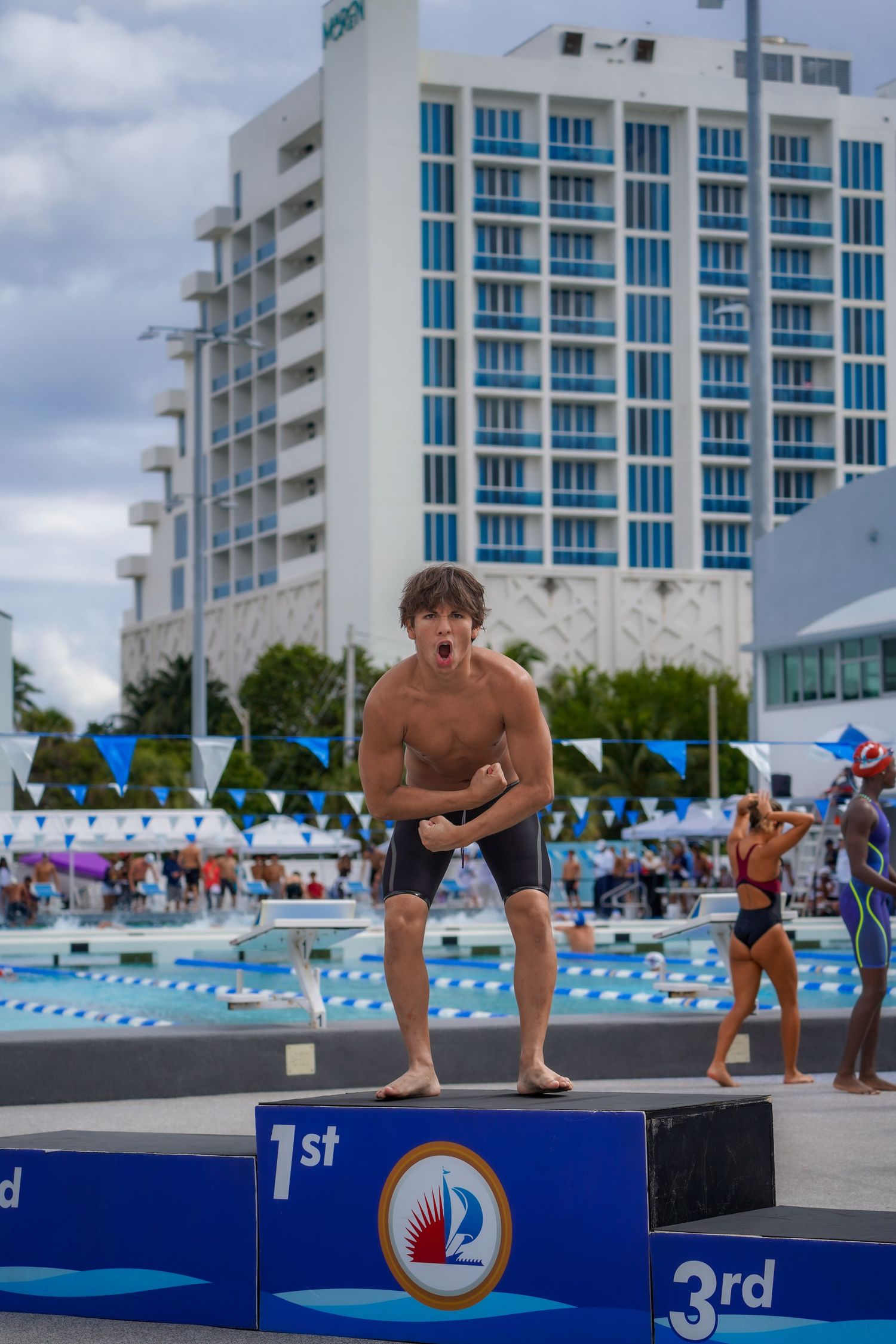 Competitors positioned on diving blocks at an outdoor swimming pool with white buildings in background.