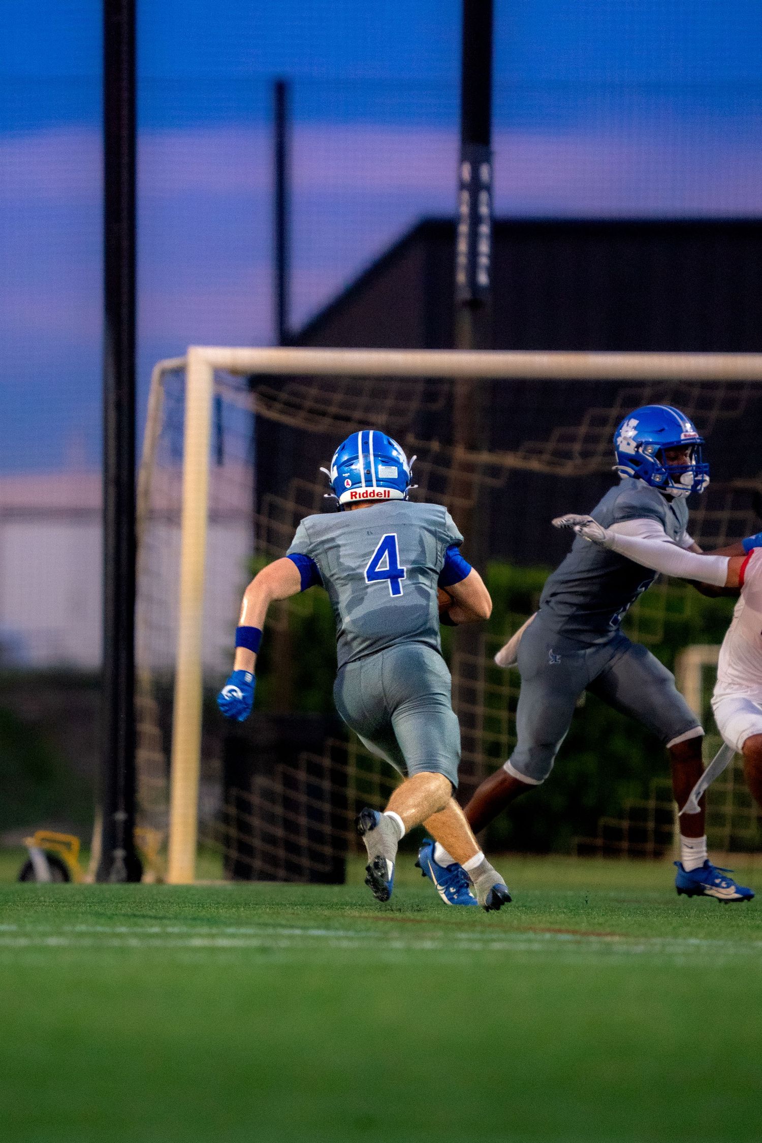 Football players execute diving plays near the goal posts at dusk.